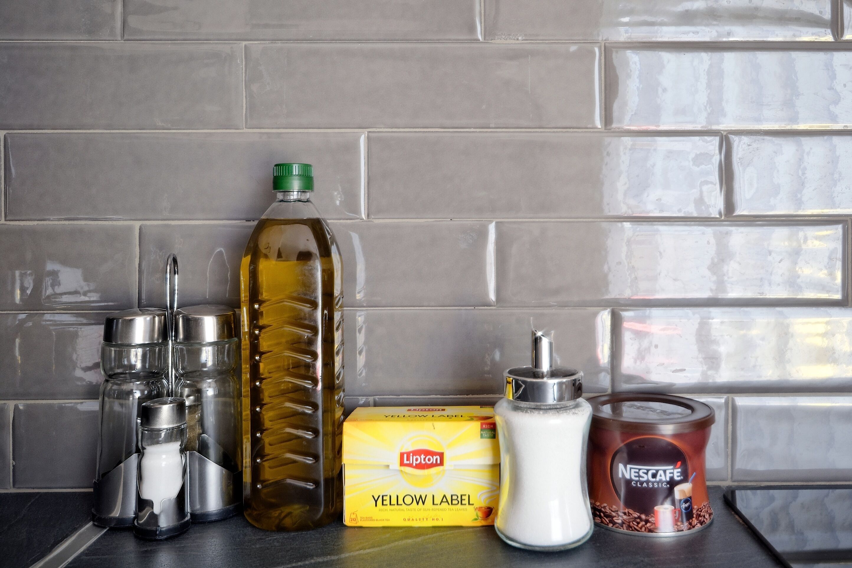 A kitchen counter with a bottle of oil, a box of Lipton tea, a Nescafe Classic coffee can, and a salt shaker.