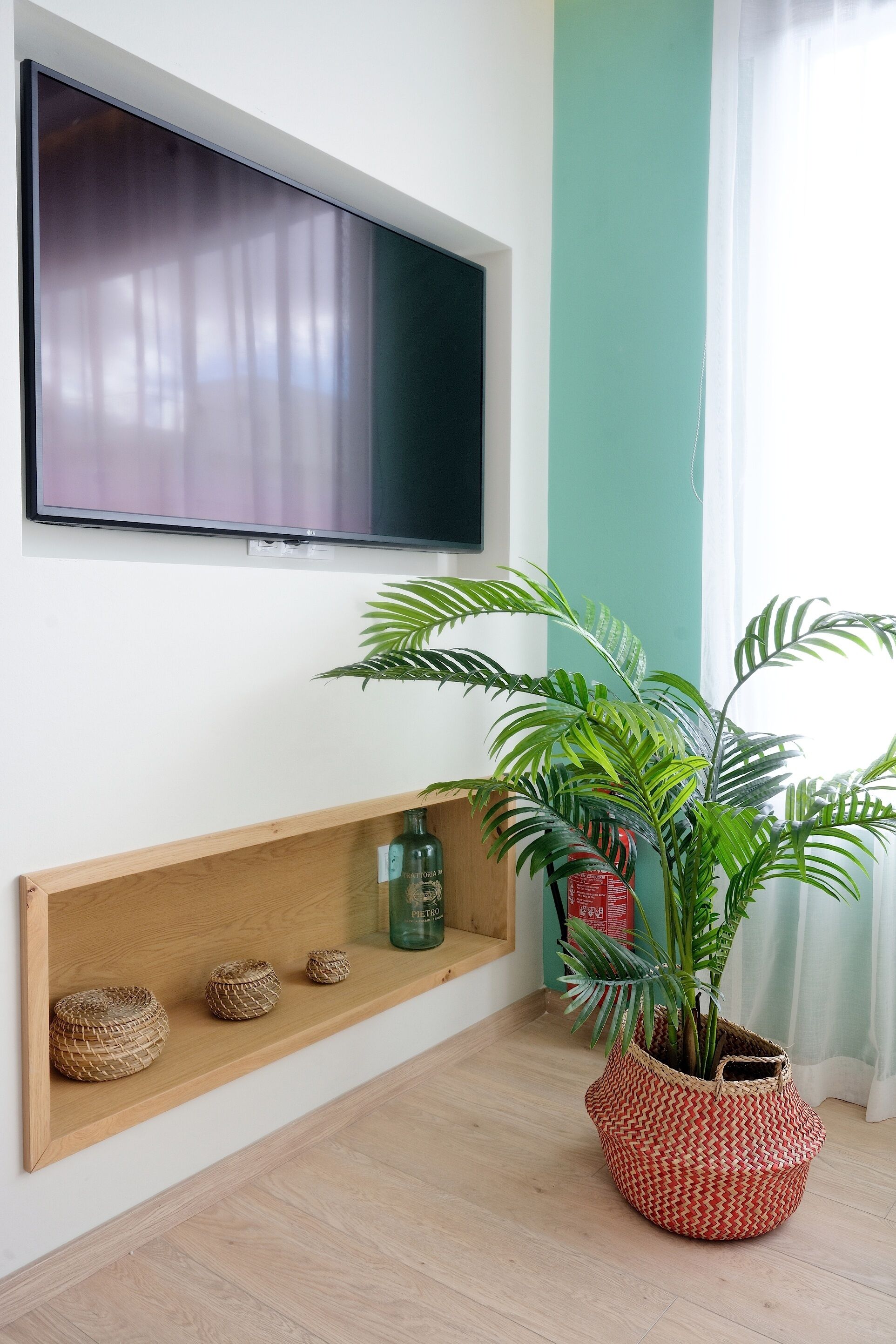 A room with a mounted TV, a wooden shelf with decorative items, and a potted plant.