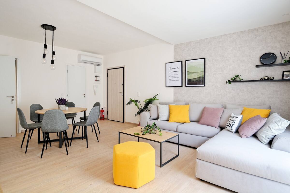 Una sala de estar moderna con un sofá gris, un otoman amarillo y una zona de comedor con sillas grises y una mesa redonda.