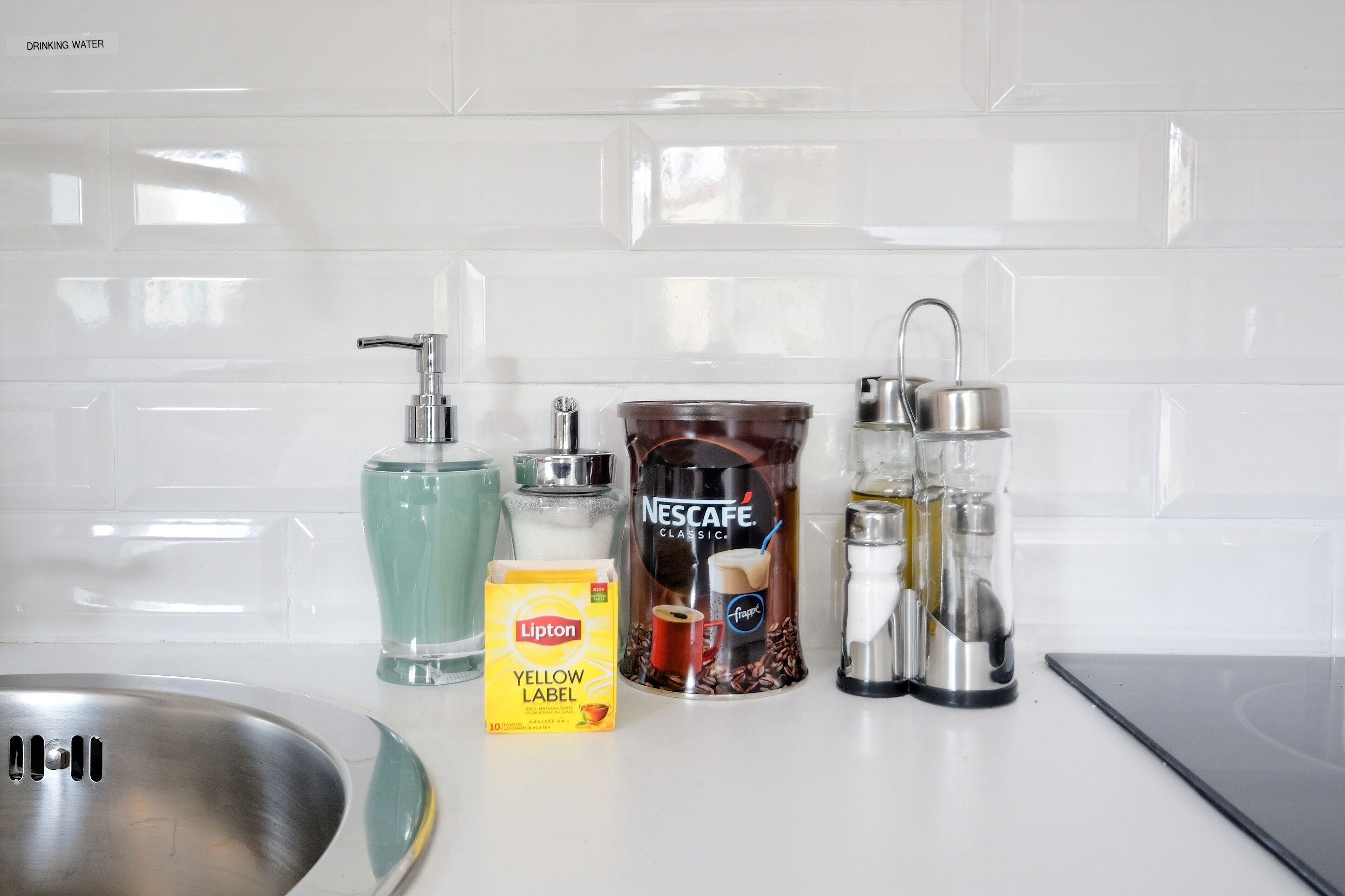 Un piano di lavoro in cucina con una macchina da caffè Nescafe Classic, una bustina di tè Lipton Yellow Label, un bicchiere d'acqua e un lavello in acciaio inossidabile.