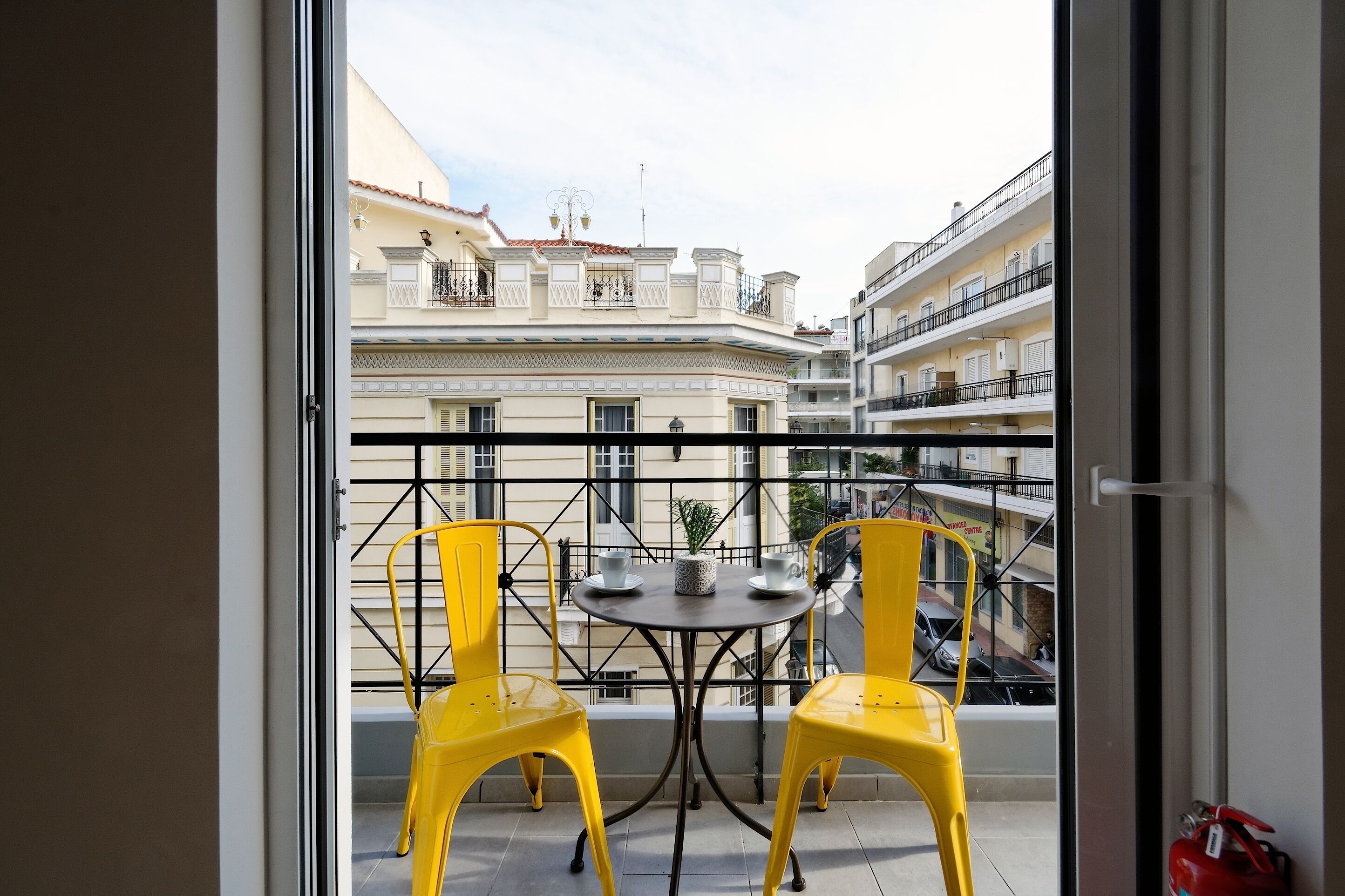 Ein Balkon mit gelben Stühlen und einem kleinen Tisch, mit Blick auf ein Wohngebiet mit Balkonen und Gebäuden.