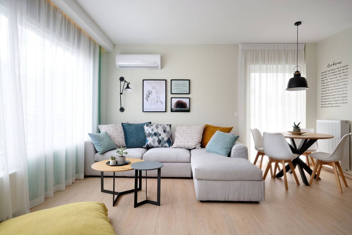 Una sala de estar moderna con un sofá, reposapiés, una mesa de centro redonda y una mesa de comedor con sillas. Hay un aire acondicionado en la pared y una lámpara colgante sobre la mesa de comedor.