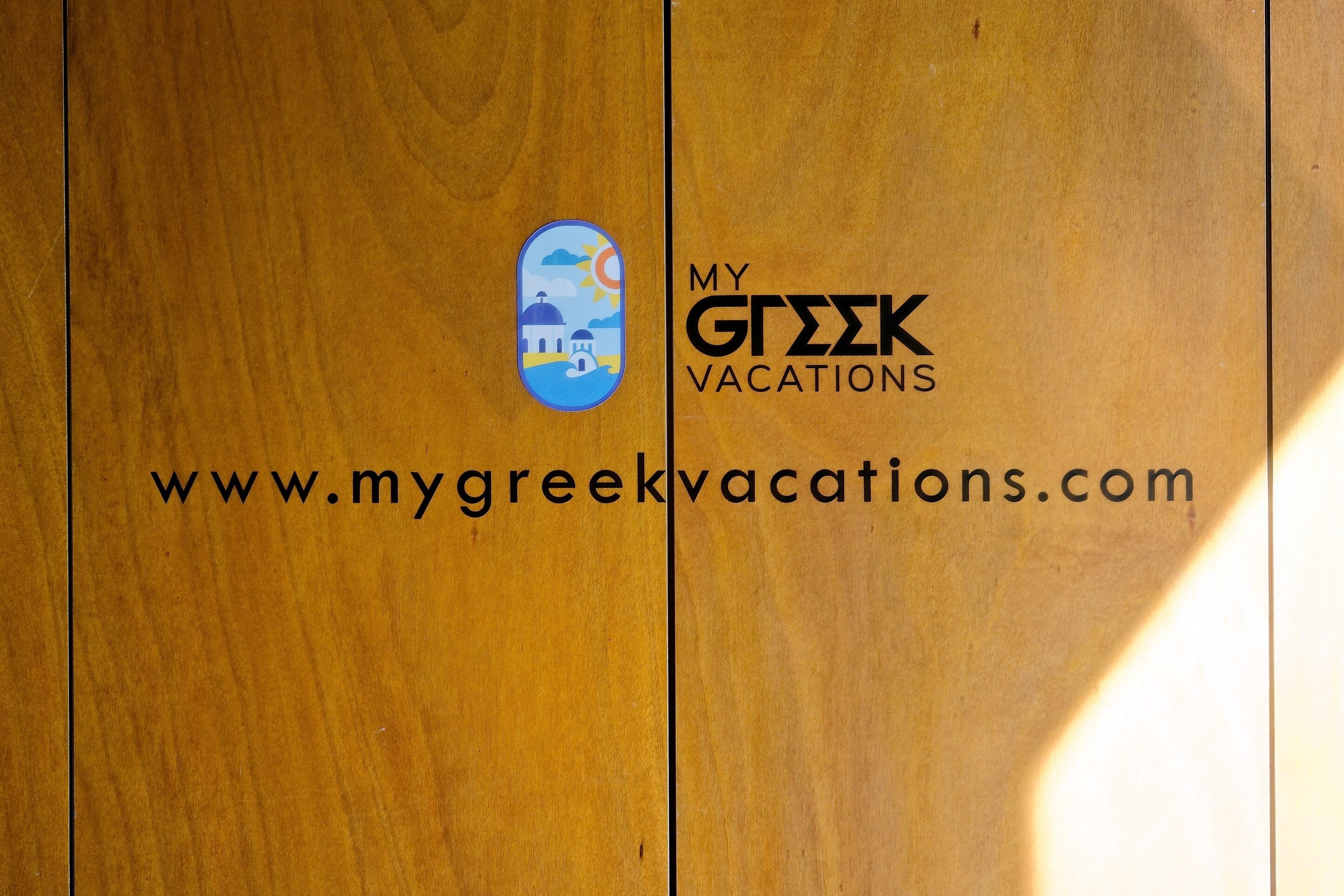 Una puerta de madera con un logotipo y el texto "MY GREEK VACATIONS" y una dirección web.