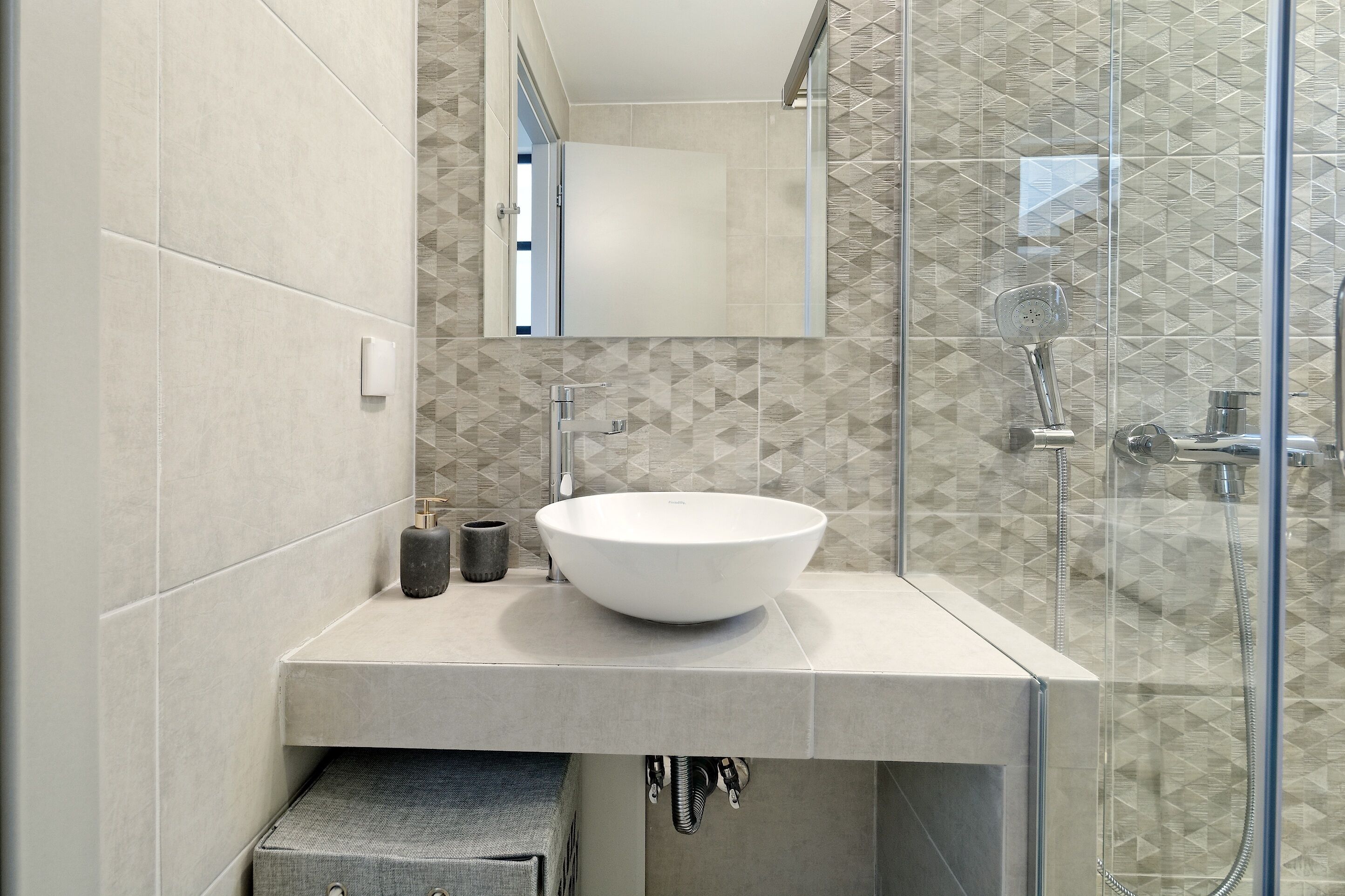 Ein modernes Badezimmer mit einem großen weißen Waschbecken, einer Duschkabine aus Glas und einer geometrischen Fliesenrückwand.