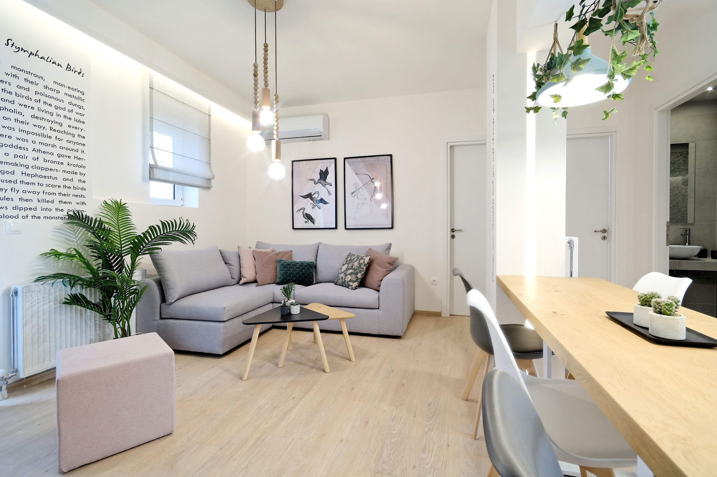 Una sala de estar moderna con un sofá gris, una mesa de comedor de madera y una pequeña mesa de centro. Hay un radiador, una planta en maceta y cuadros en la pared.