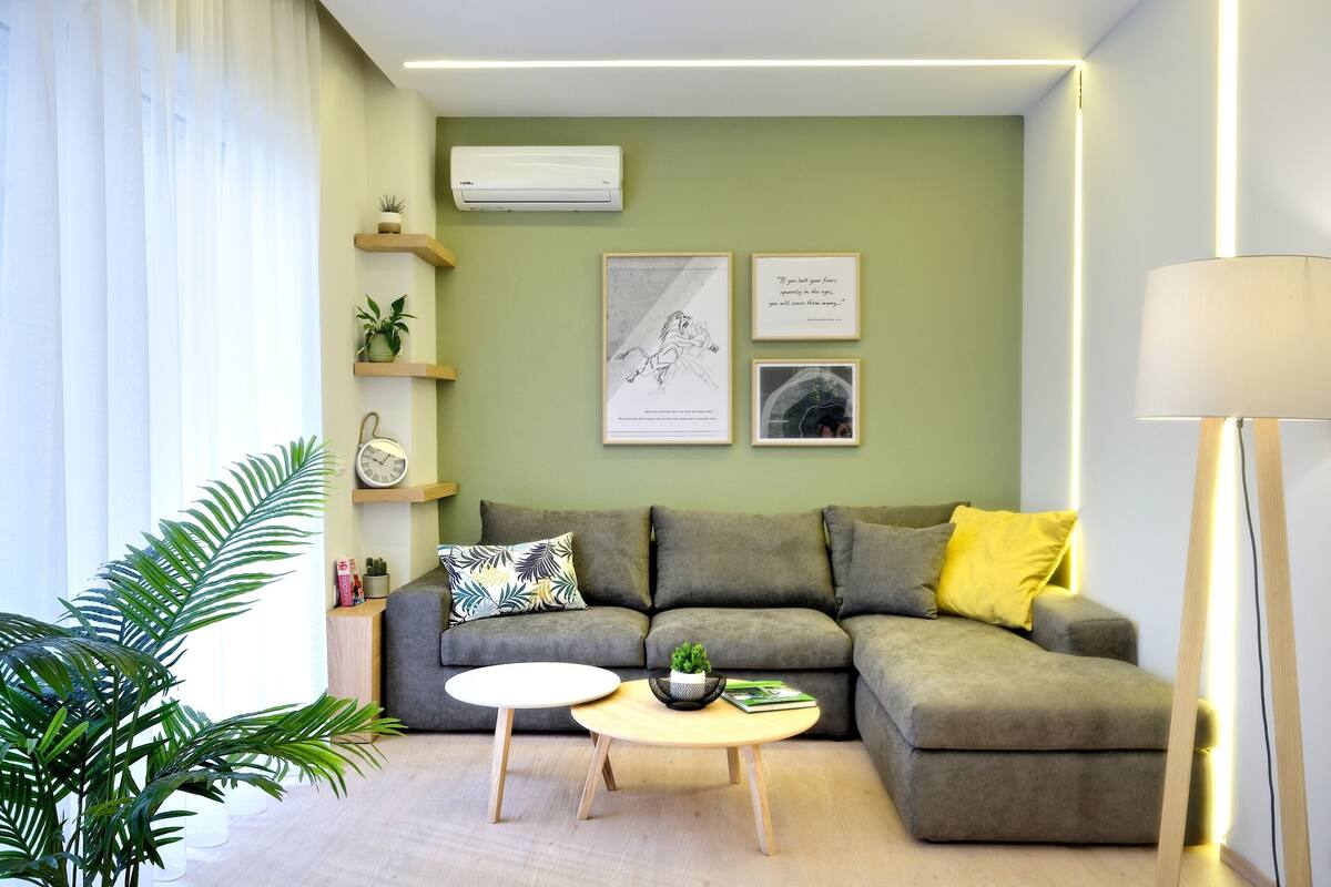 Una sala de estar moderna con un sofá gris, una mesita de centro redonda y una pequeña mesita auxiliar. Hay estantes en la pared con plantas y obras de arte enmarcadas.
