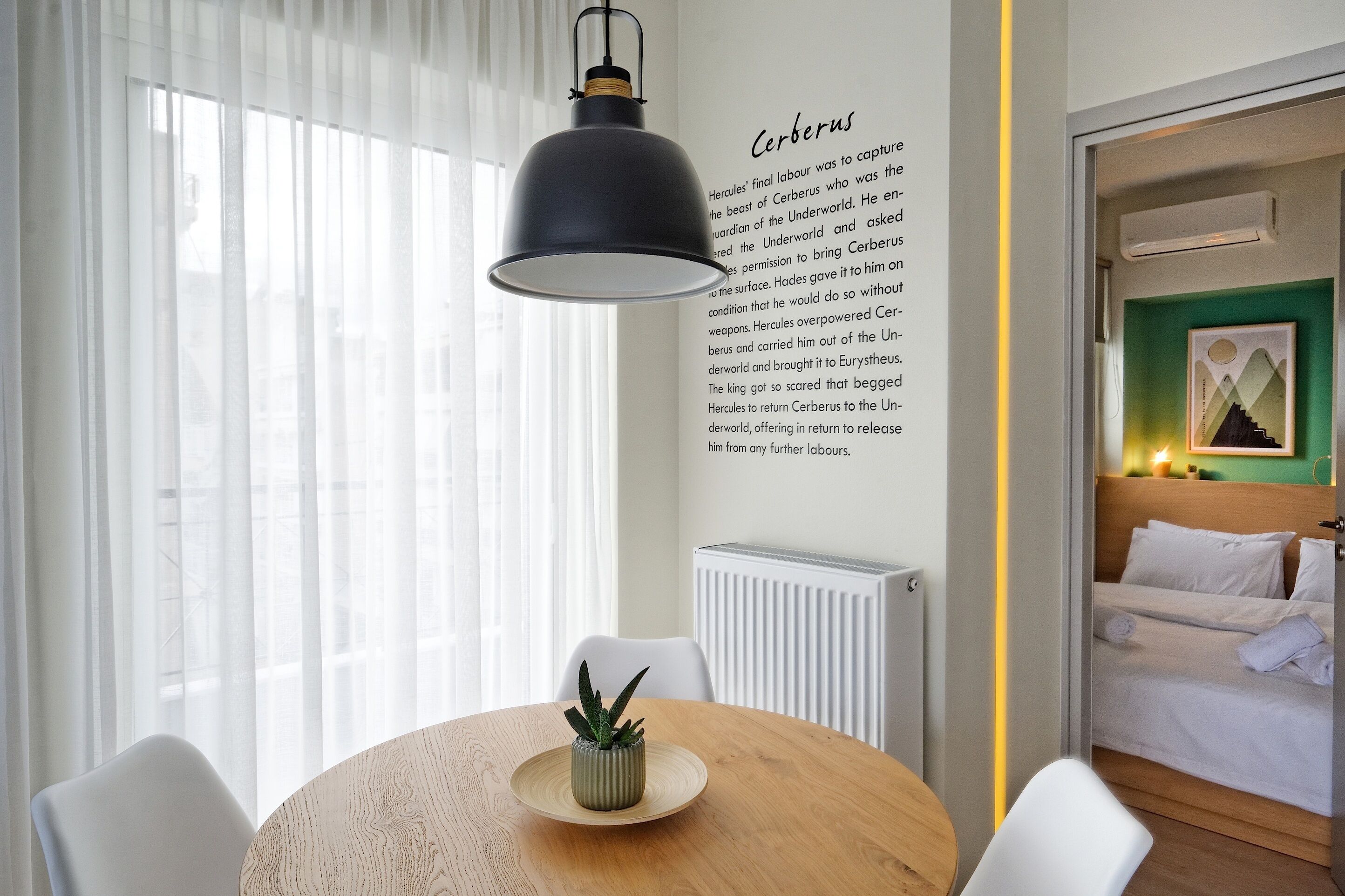 A hotel room with a round wooden table, a potted plant, white chairs, a bed with white bedding, and a wall with a text about Cerberus.