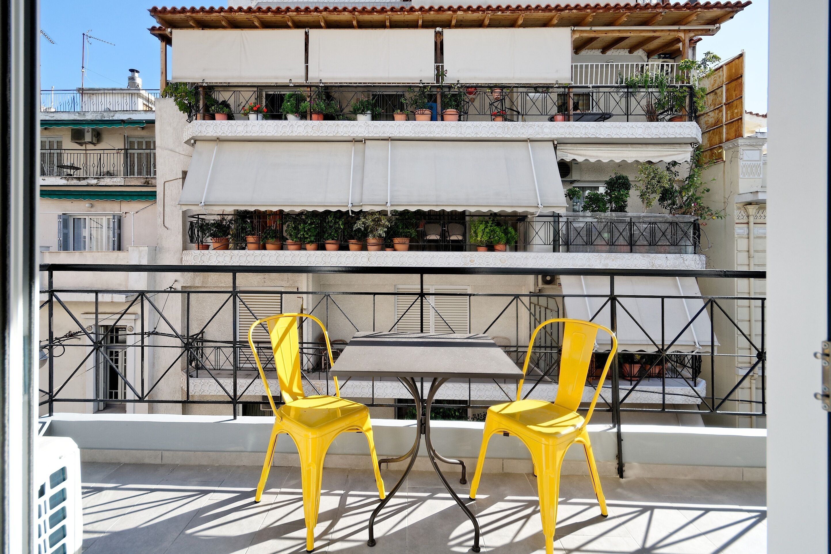 A balcony with two yellow chairs and a table, overlooking a building with multiple balconies and potted plants.