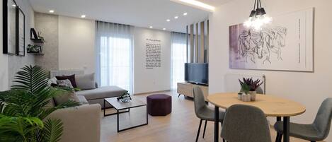 Una sala de estar moderna con un sofá, una mesa de centro y un comedor con sillas. Hay un televisor y obras de arte en la pared.