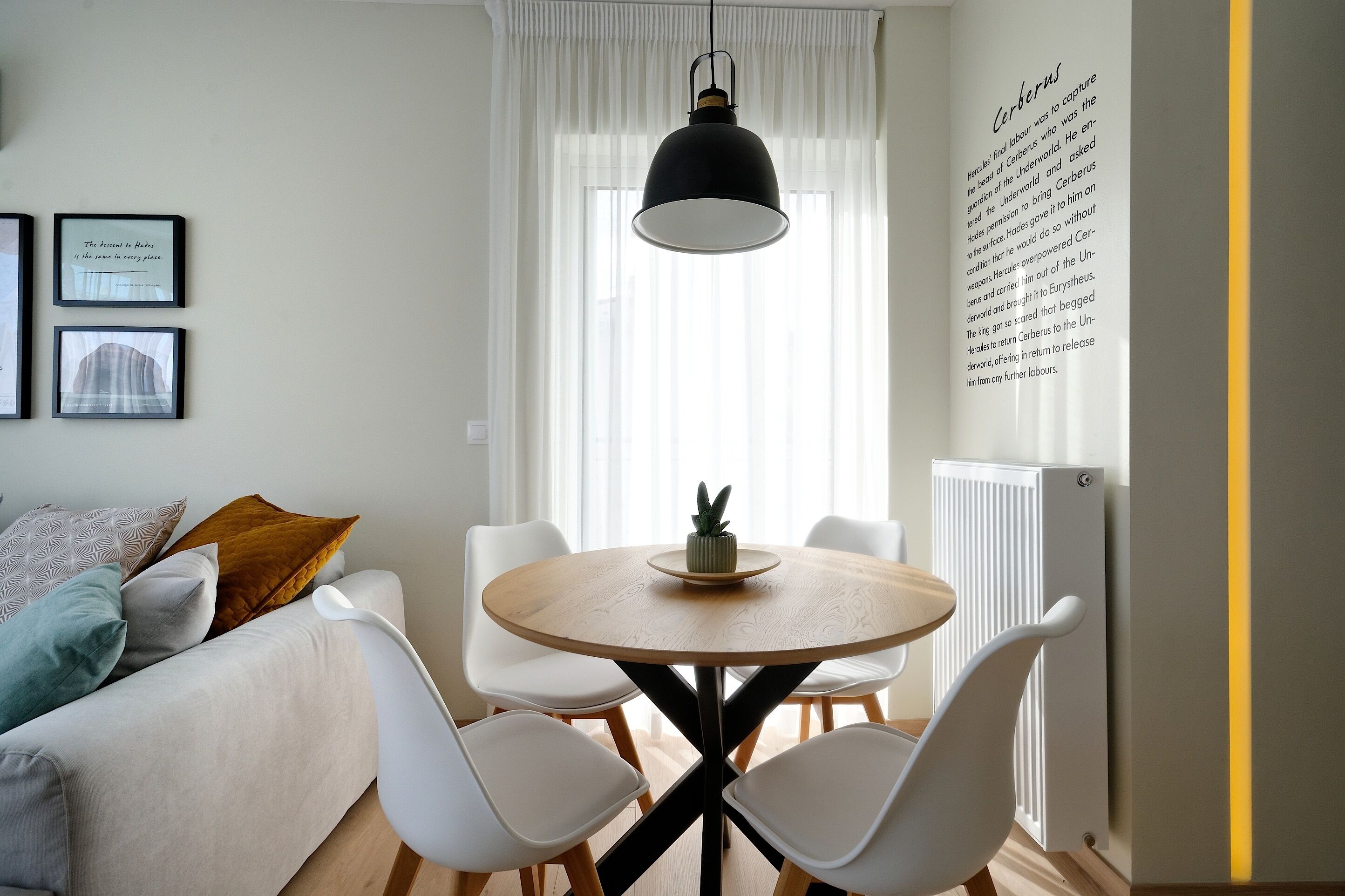 A modern living room with a round wooden table, white chairs, a black hanging lamp, and a radiator. There are framed pictures on the wall and a window with white curtains.