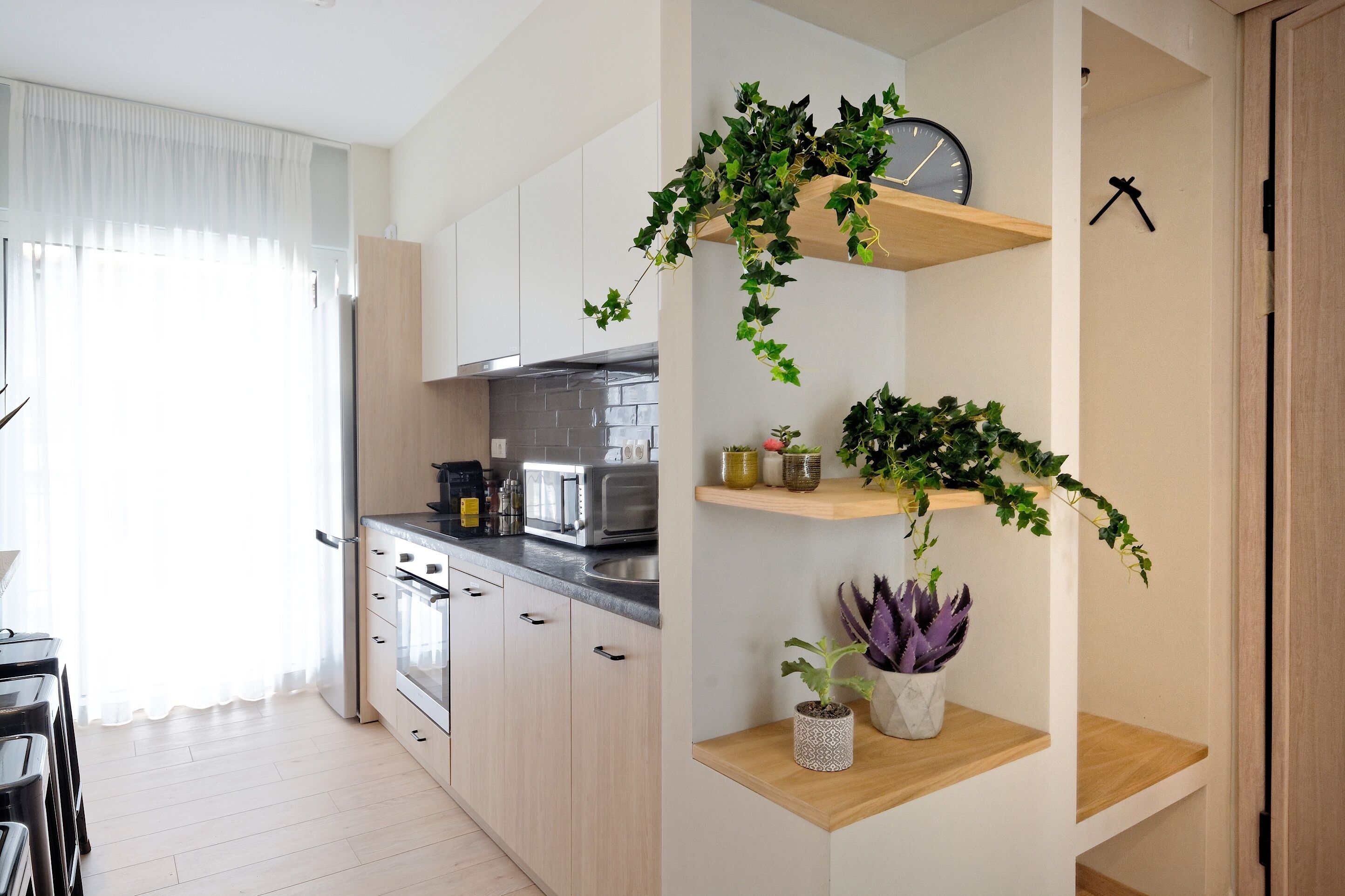 A modern kitchen with white cabinets, a microwave, and a sink. There are wooden shelves with plants and a clock.