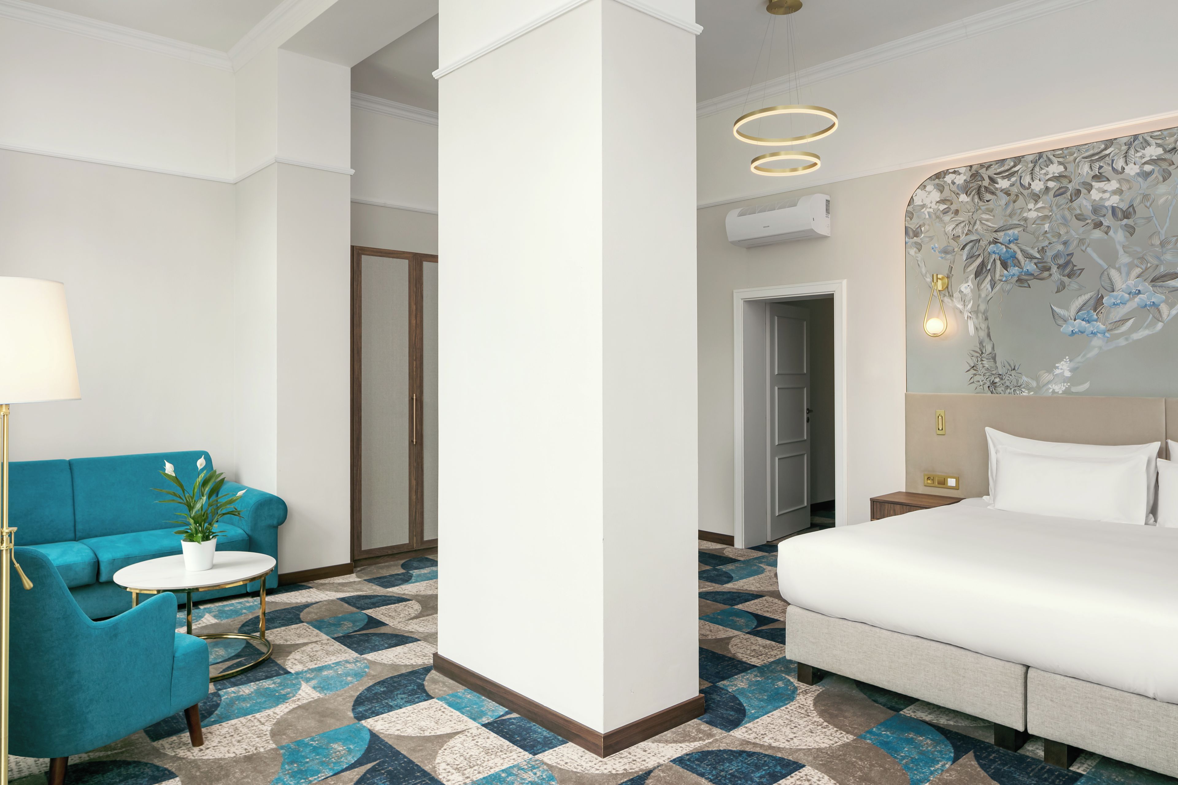Quarto de hotel com cama, sofá azul, mesinha com abajur e piso estampado.