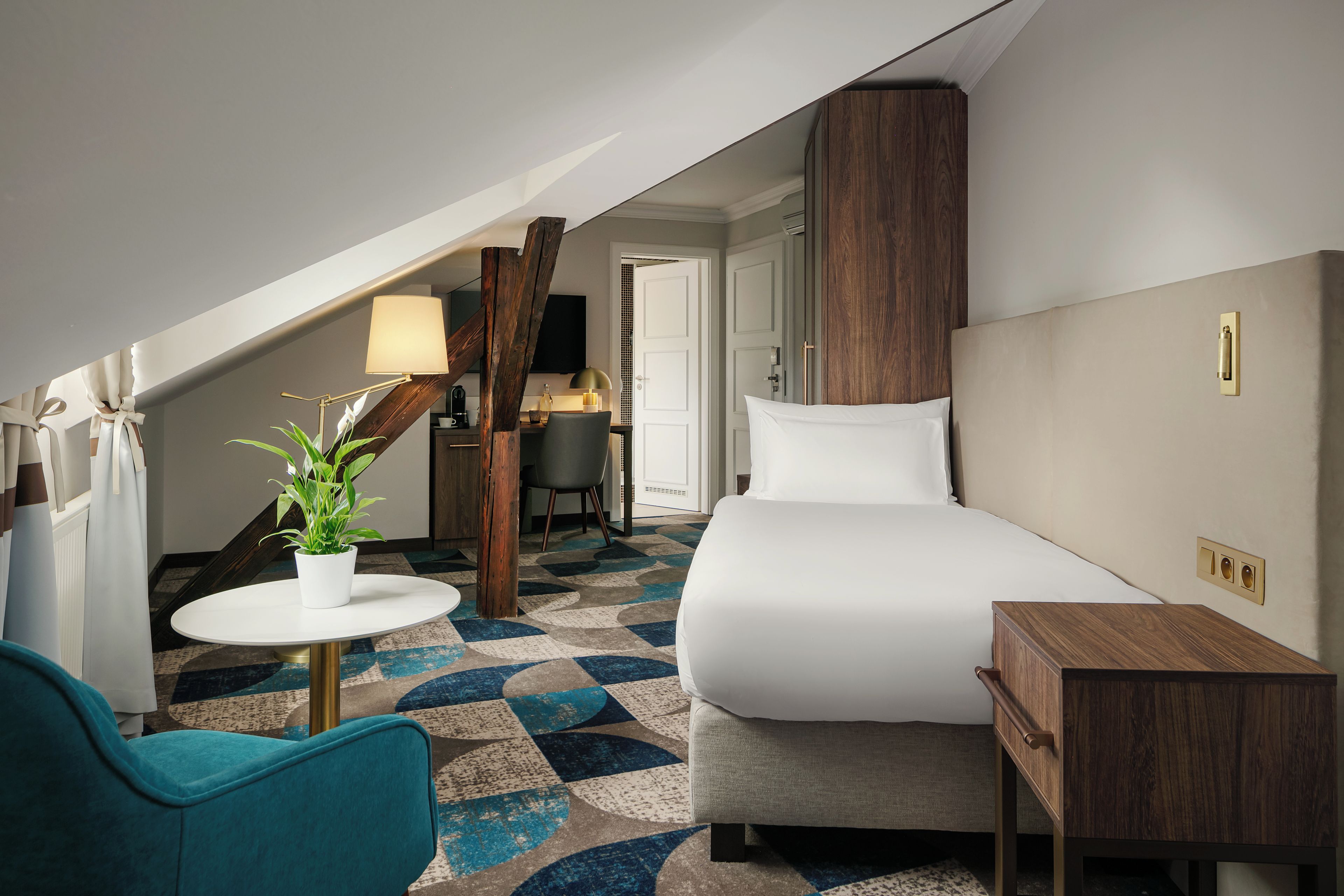 A hotel room with a bed, a small table, a chair, a potted plant, and a lamp.