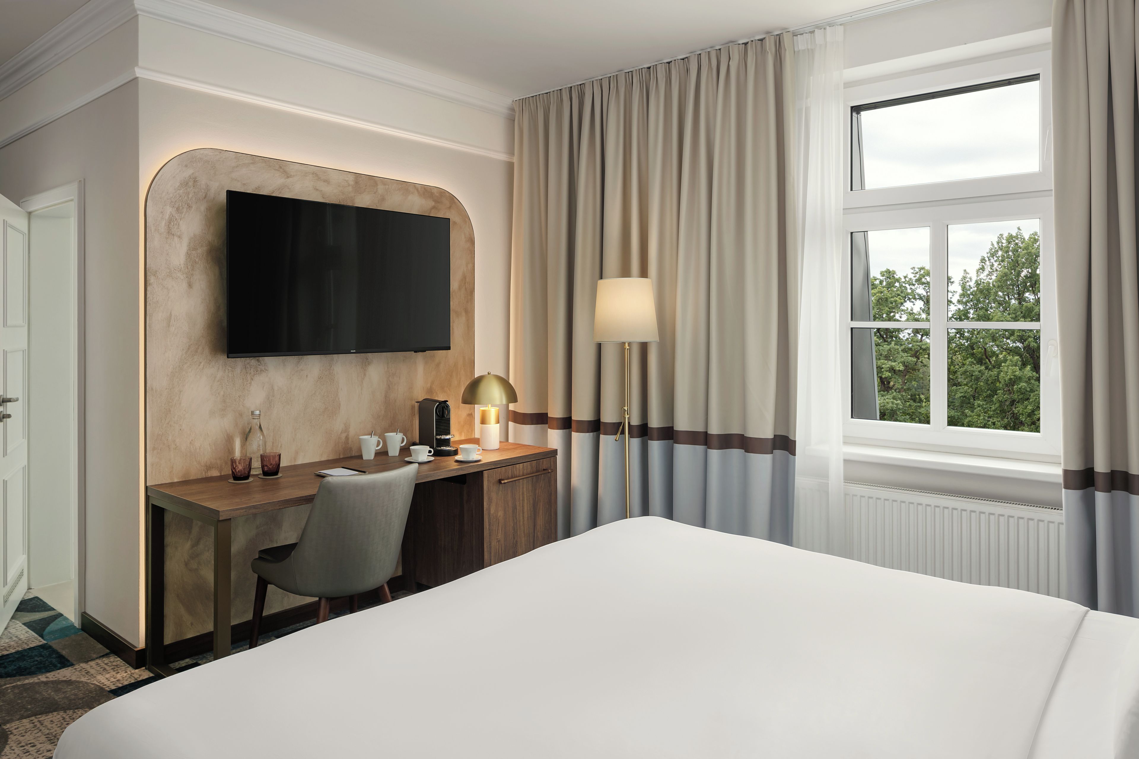 Ein Hotelzimmer mit einem Bett, einem Schreibtisch, einem Flachbildfernseher, einer Lampe und einem Fenster mit Vorhängen.