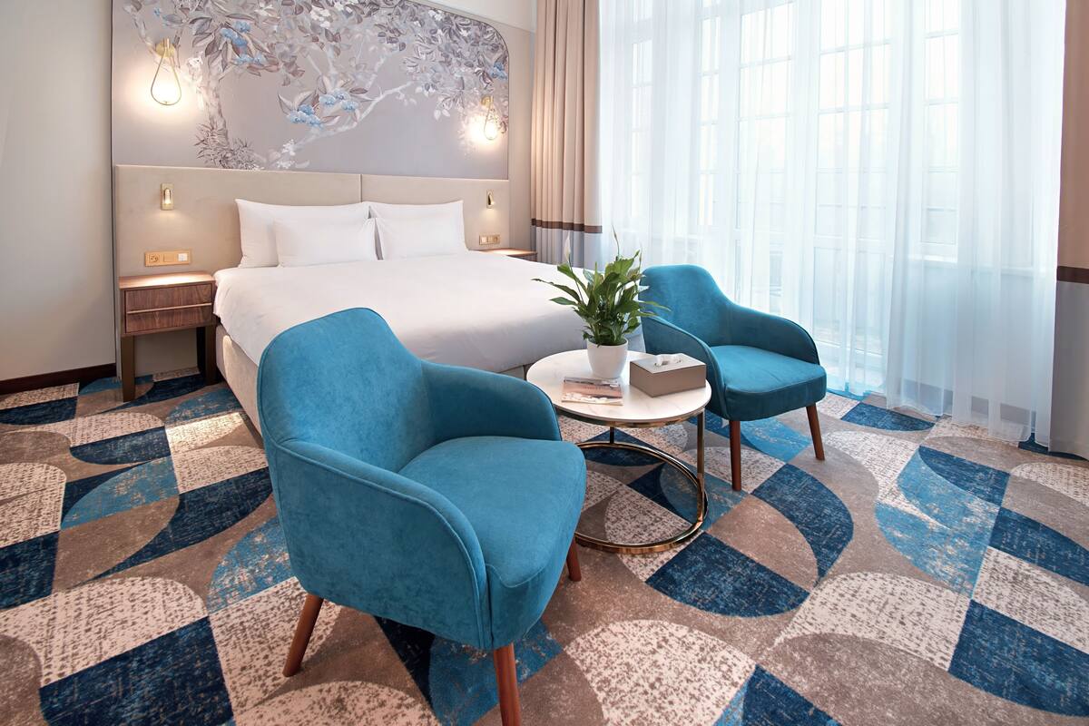 A hotel room with a large bed, two blue armchairs, a small round table, and a potted plant.