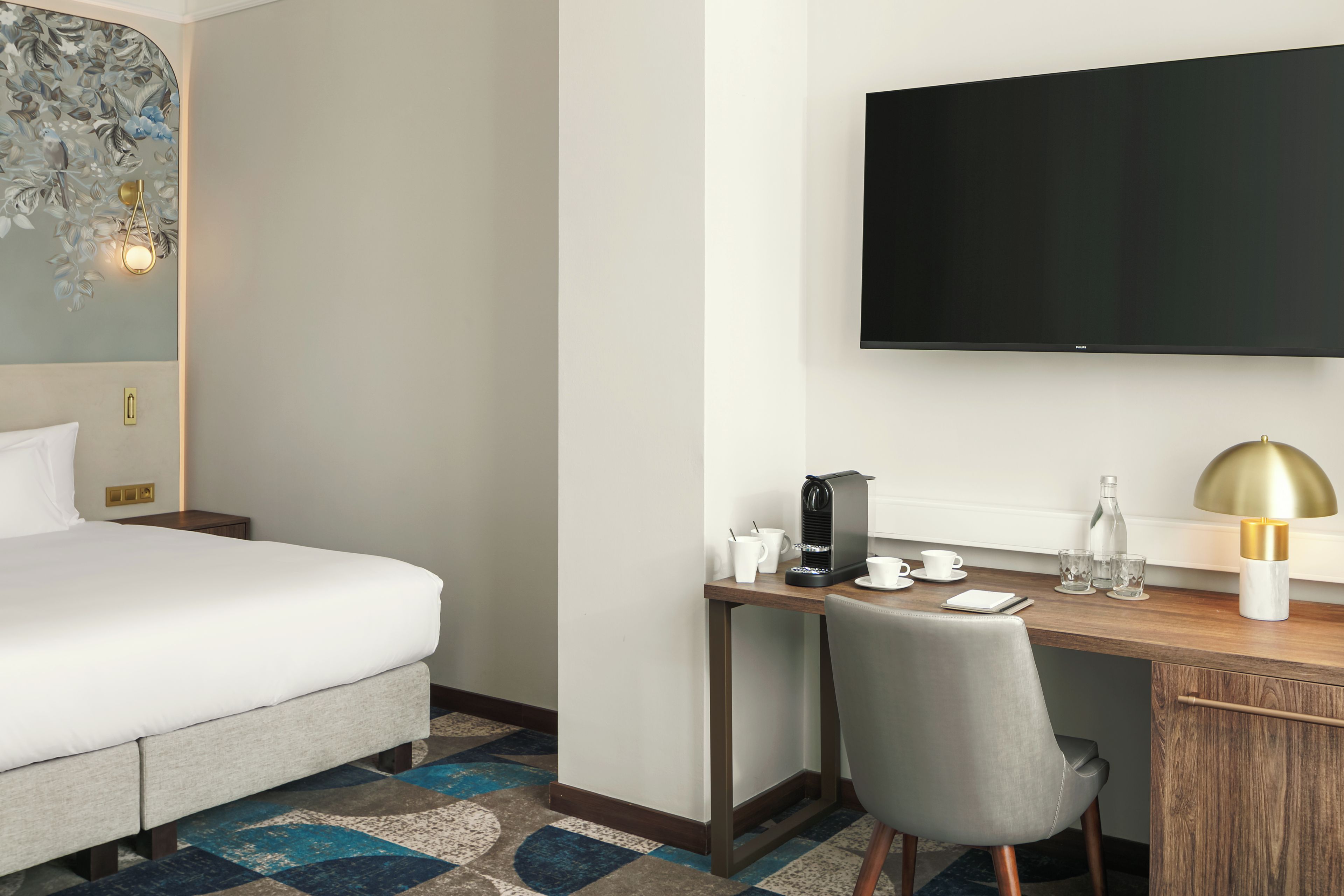 A hotel room with a bed, a desk with a coffee machine, a TV, and a chair.