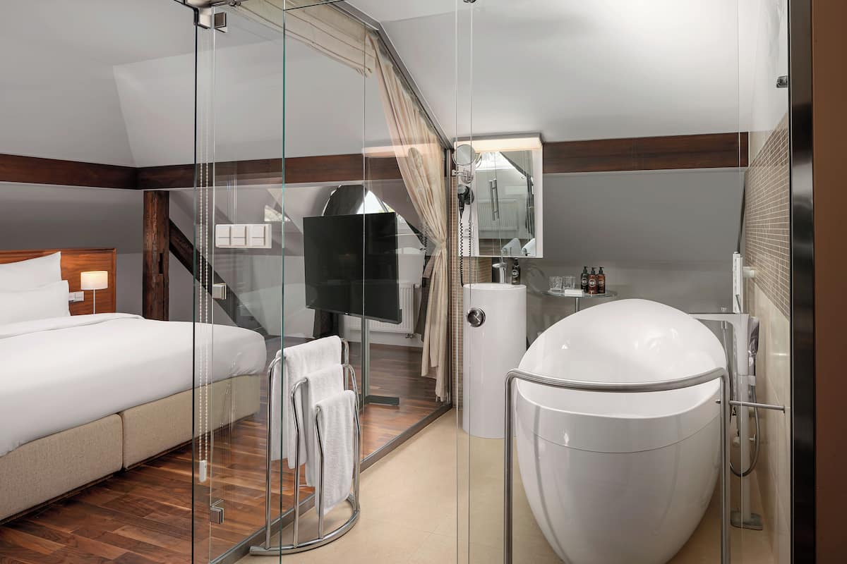 A modern hotel room with a glass-enclosed shower, a large bed, a vanity with a mirror, and a toilet.