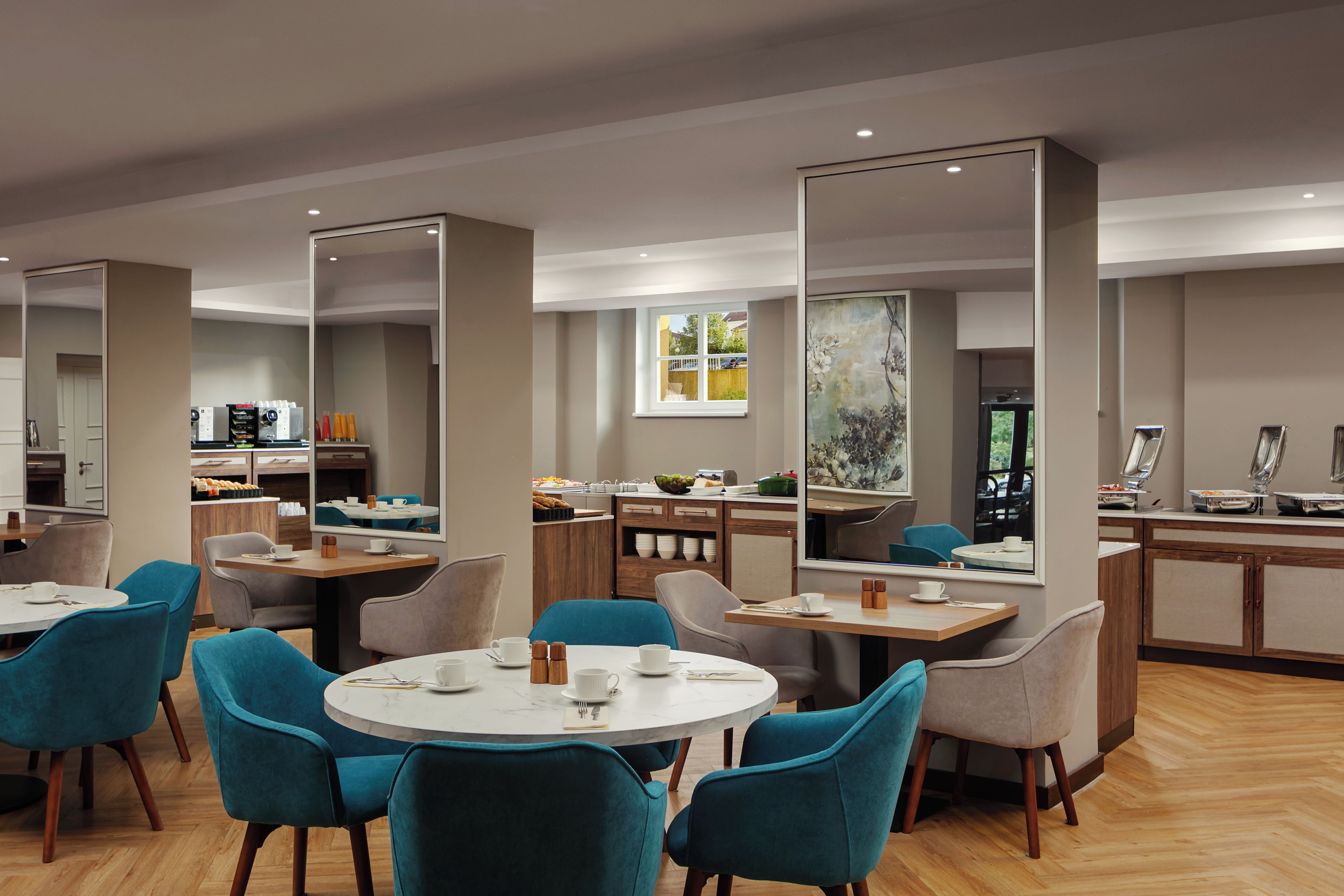Un comedor moderno con mesas redondas y sillas color turquesa, una cocina al fondo y un ventanal con vistas.