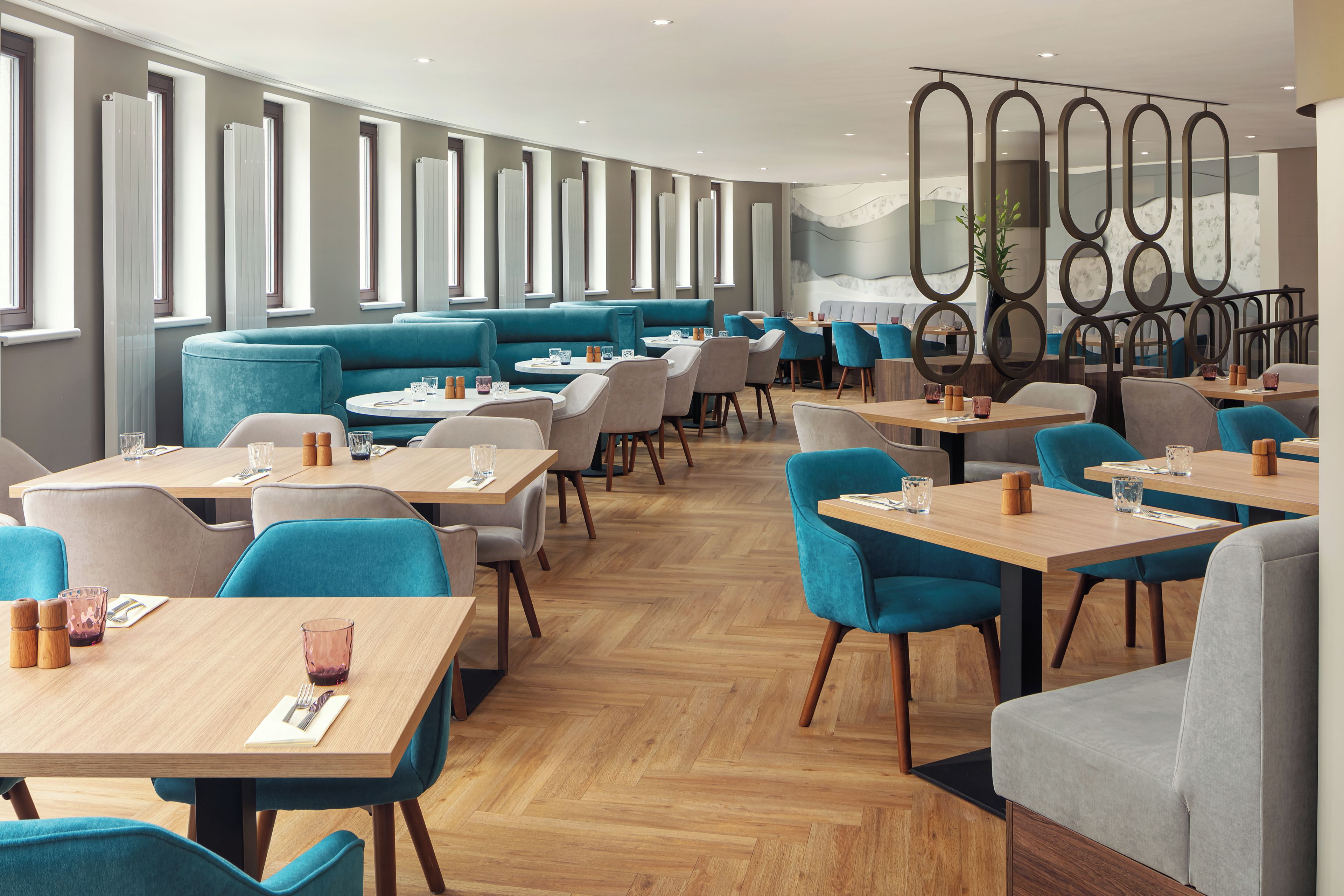 A modern restaurant interior with high tables and chairs, a herringbone wood floor, and a seating area with blue upholstered sofas.