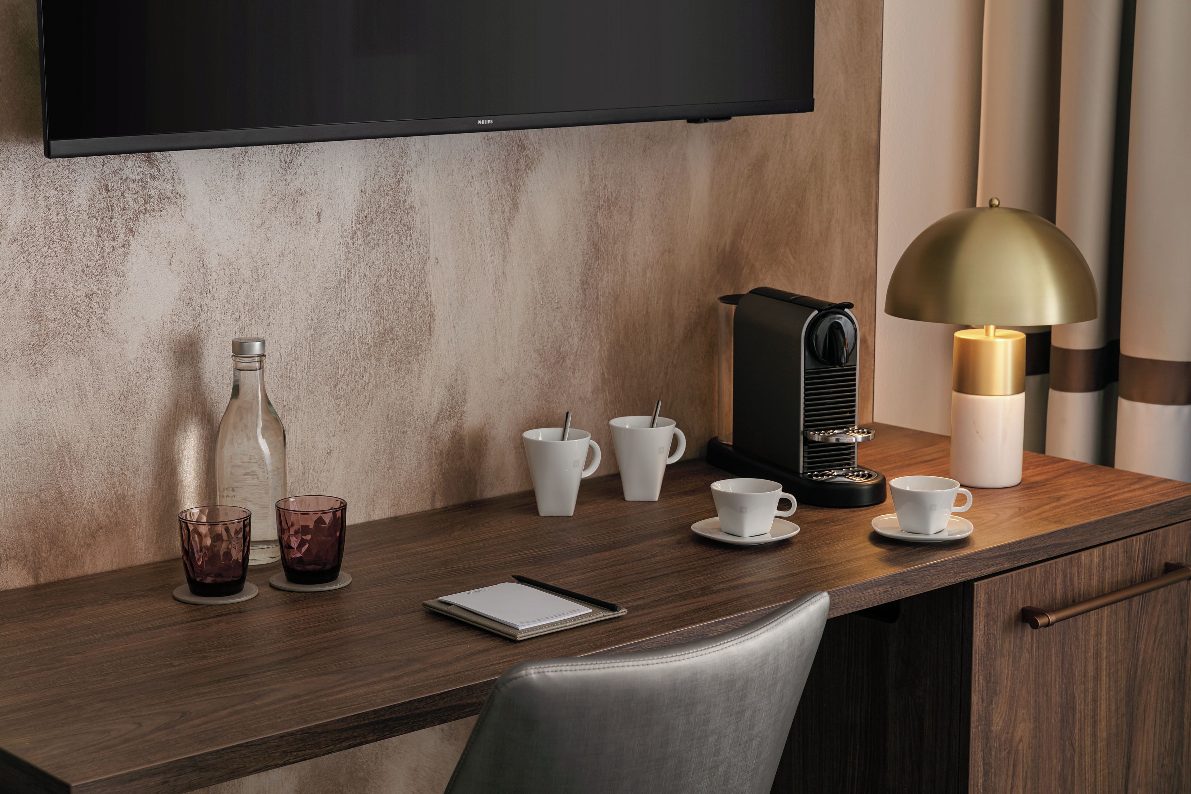 A hotel desk with a flat-screen TV, a coffee machine, and various cups and glasses.