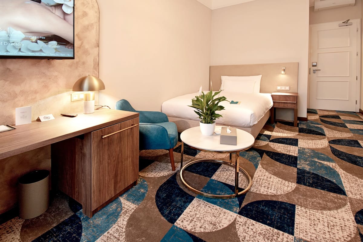 A hotel room with a bed, bedside table, lamp, chair, small table with a plant, and a patterned carpet.