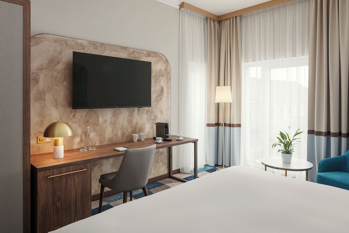 A hotel room with a flat-screen TV, a desk with a lamp, a coffee machine, a chair, a bed, and a small table with a plant.
