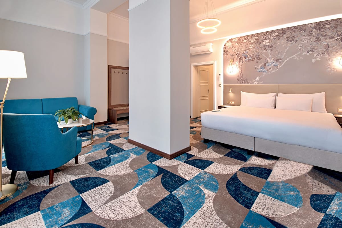 A hotel room with a bed, a blue armchair, a small table with a plant, and a patterned carpet.