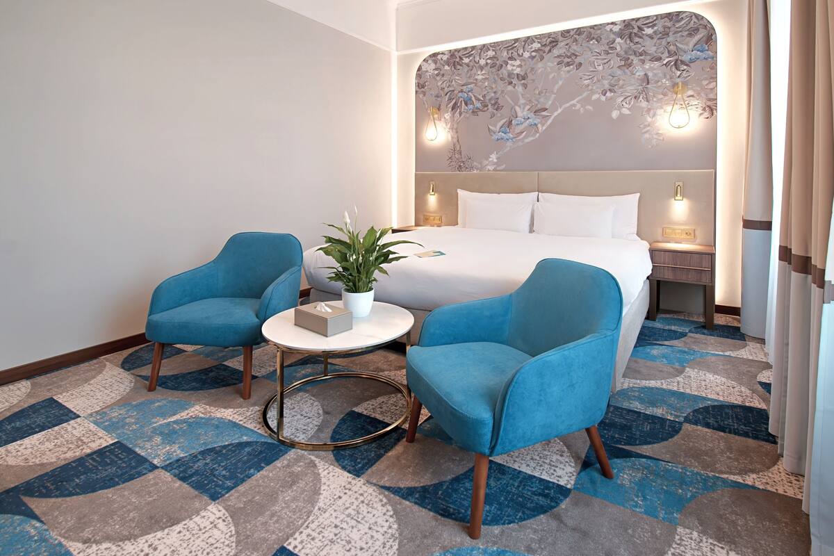 A hotel room with a bed, two blue armchairs, a small round table with a plant, and a decorative wall mural.