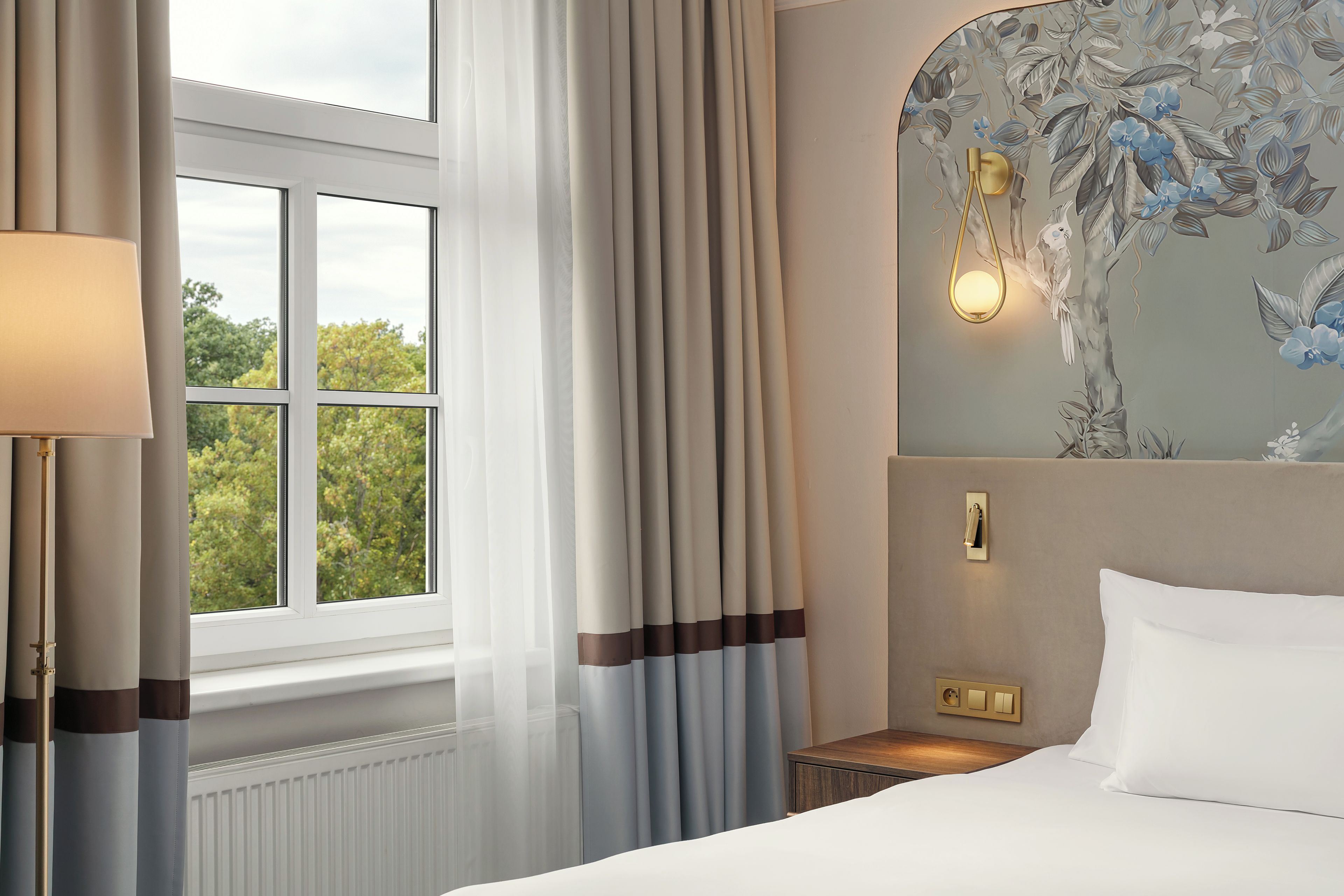 Habitación de hotel con cama, mesita de noche, lámpara, ventana con cortinas y un mural floral en la pared.
