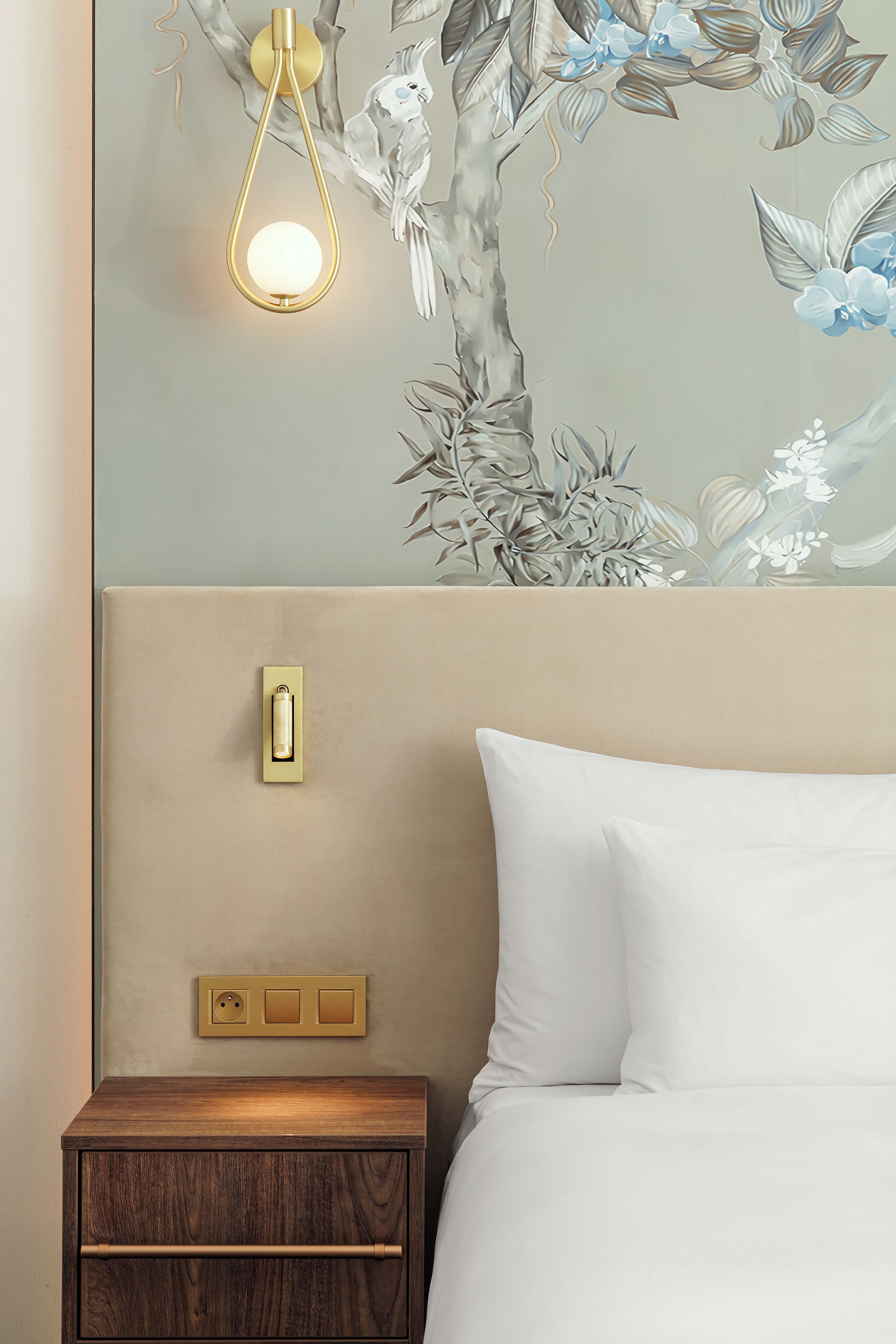 A hotel room with a bed, a nightstand, a wall-mounted light, and a power outlet.