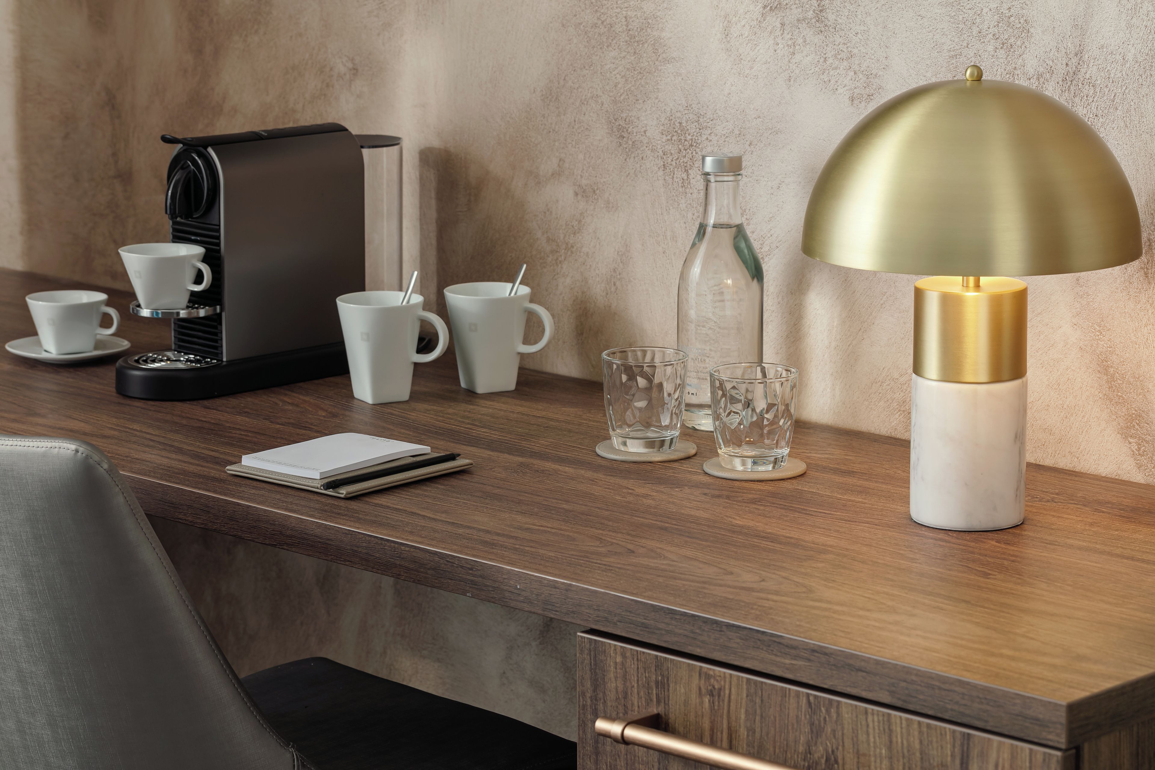 A modern hotel desk with a coffee maker, multiple cups, a water bottle, a glass, and a lamp.