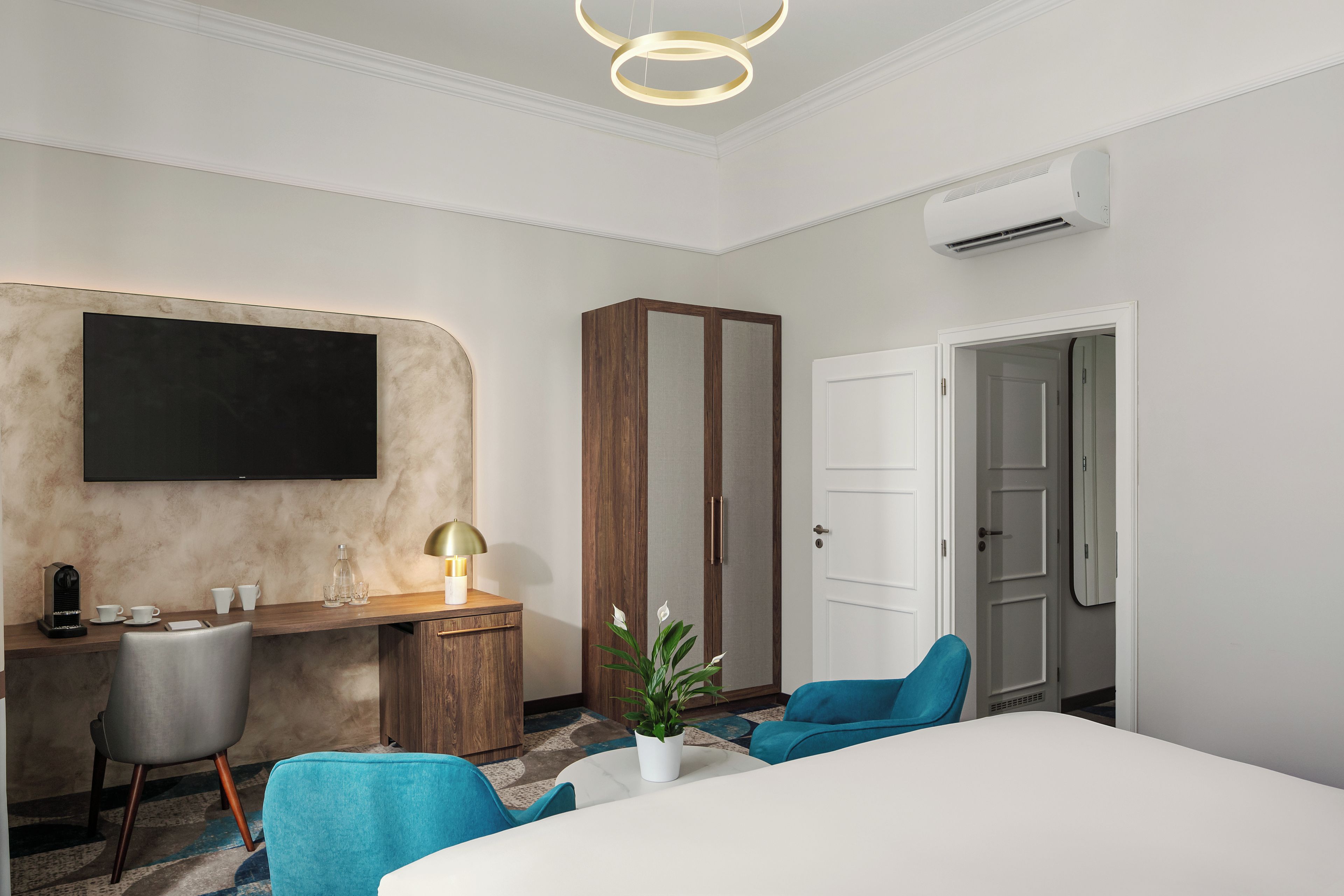 A hotel room with a bed, a desk, a chair, a television, a wardrobe, and an air conditioning unit.