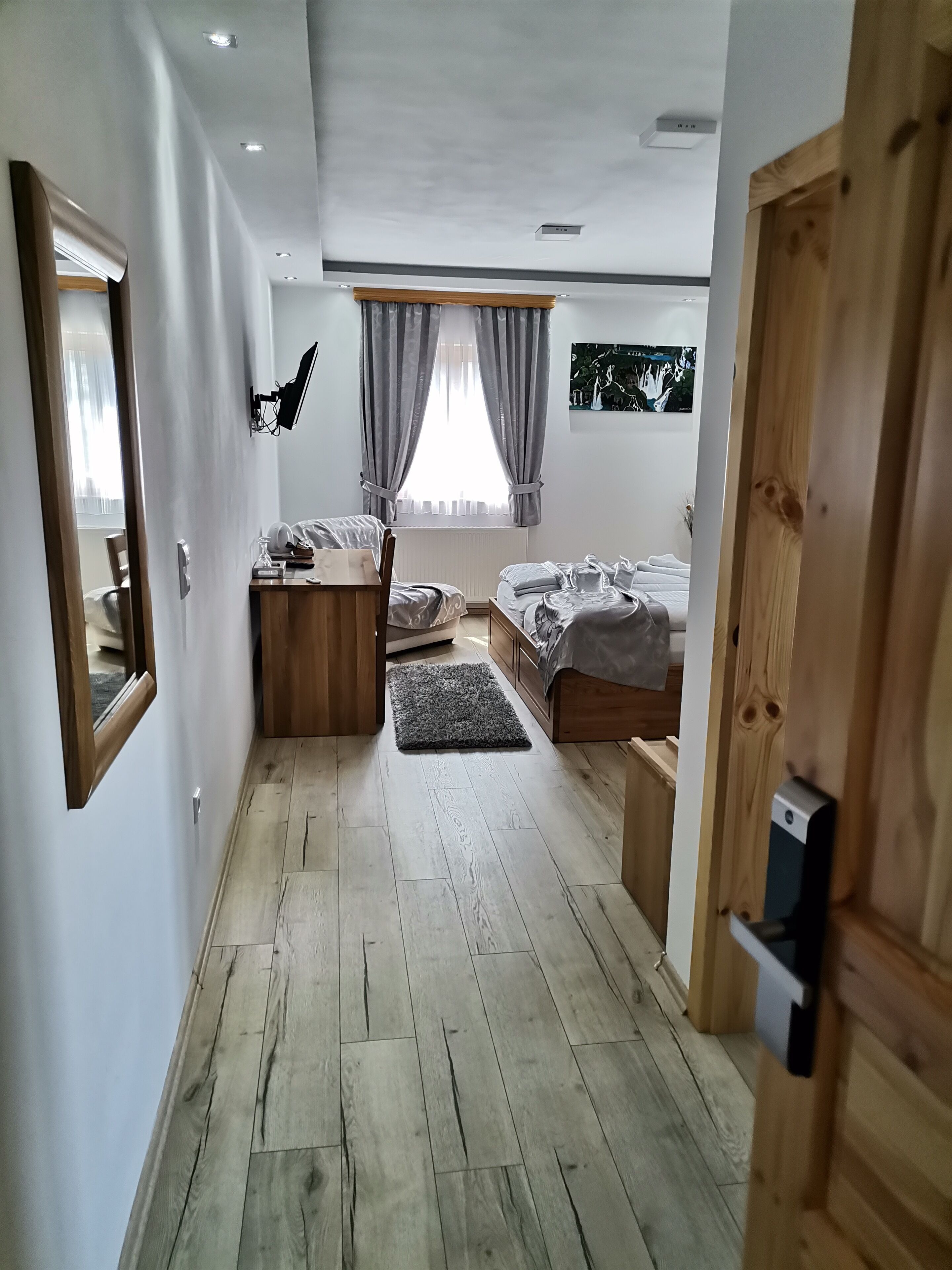 Een moderne slaapkamer met houten vloer, een bed met grijs dekbedovertrek, een bureau met computer, een raam met gordijnen en een wandgemonteerde televisie.