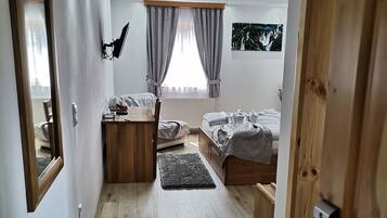 Een moderne slaapkamer met houten vloer, een bed met grijs dekbedovertrek, een bureau met computer, een raam met gordijnen en een wandgemonteerde televisie.