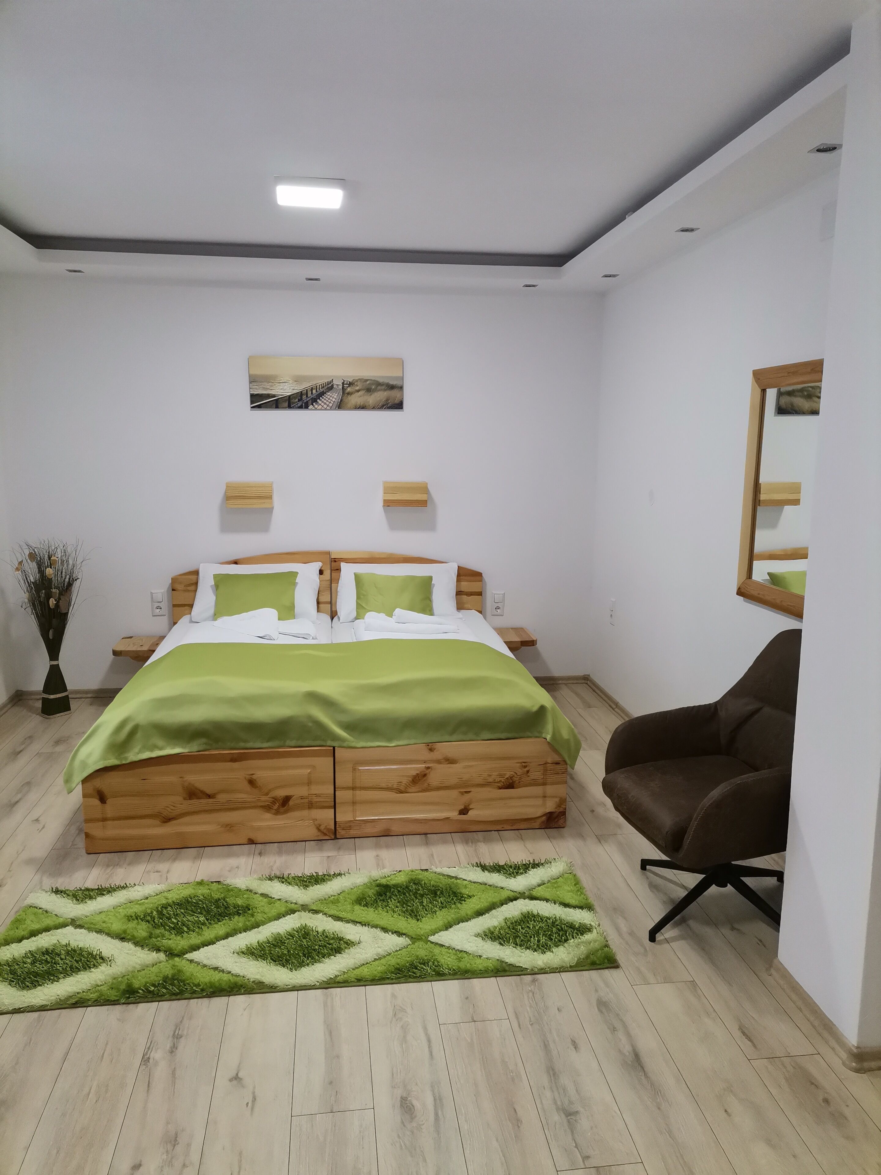 Quarto com cama de madeira, roupa de cama verde, uma pequena escrivaninha, uma cadeira, um espelho e um quadro na parede.