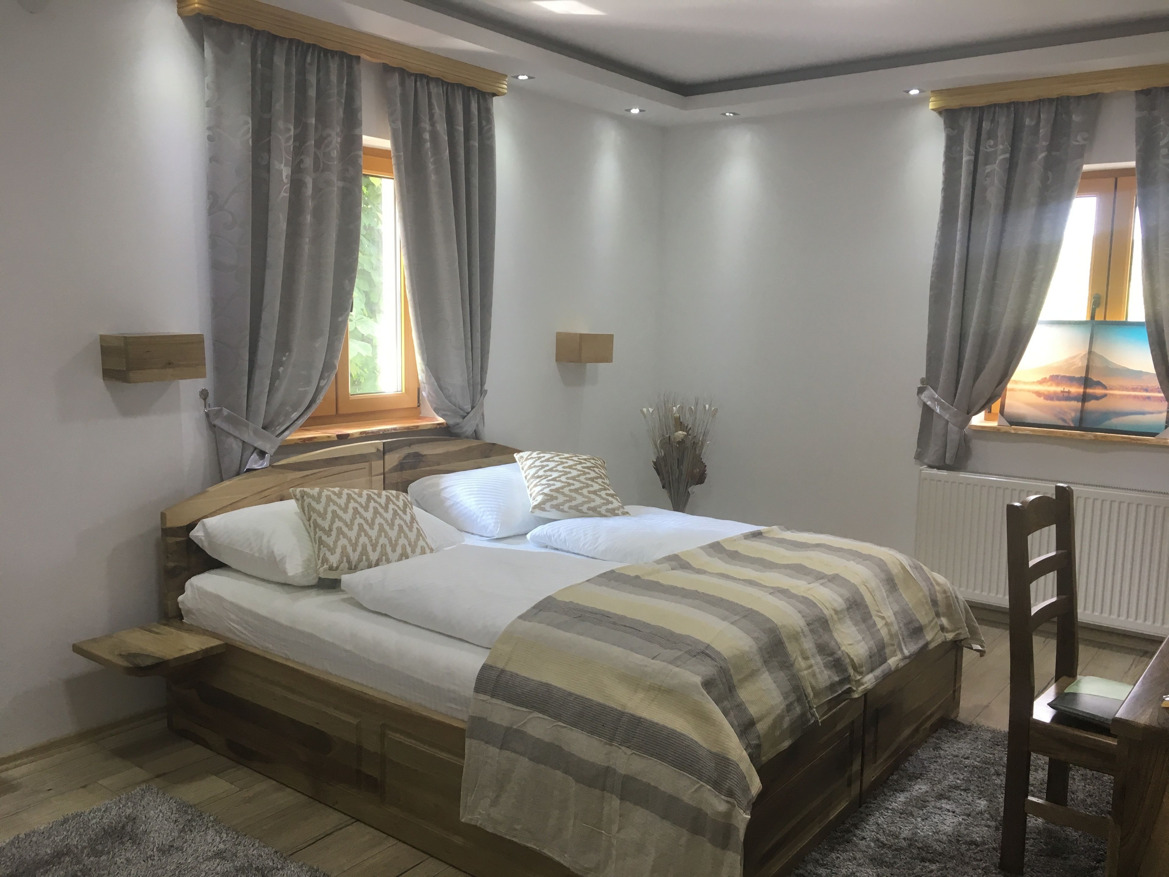 A bedroom with a wooden bed, grey curtains, a window with a view, a bedside table, and a radiator.