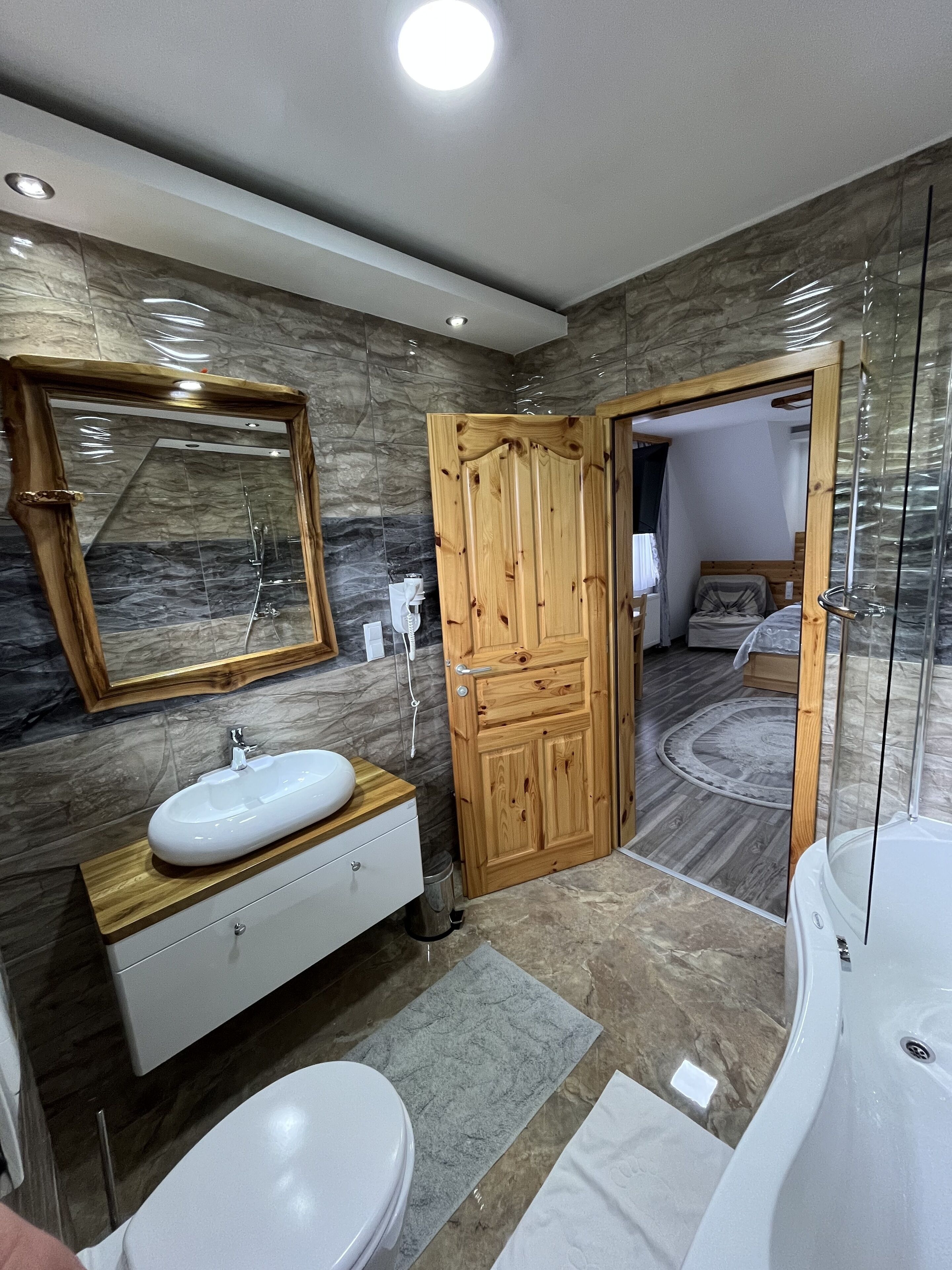 A modern bathroom with a marble wall, wooden vanity, white sink, and a glass shower enclosure.