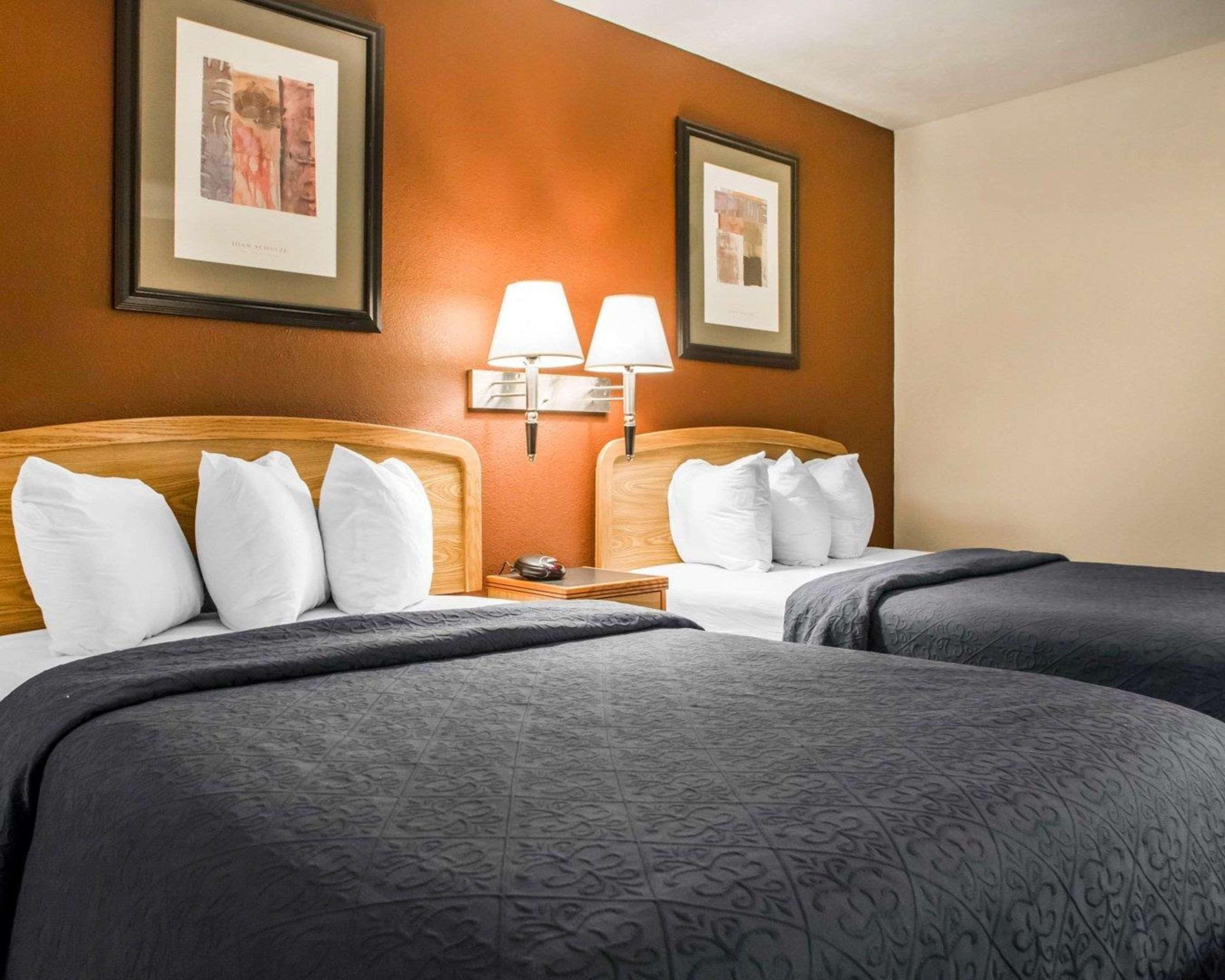 Habitación de hotel con dos camas, cada una con almohadas blancas y ropa de cama oscura. Hay dos cuadros en la pared, una mesita de noche con una lámpara y un reloj.