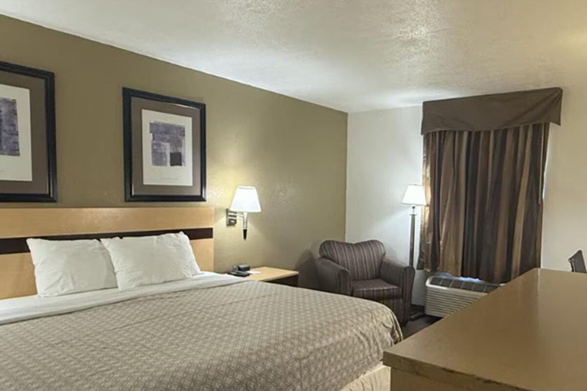 A hotel room with a bed, two framed pictures on the wall, a chair, a desk, and a window with curtains.
