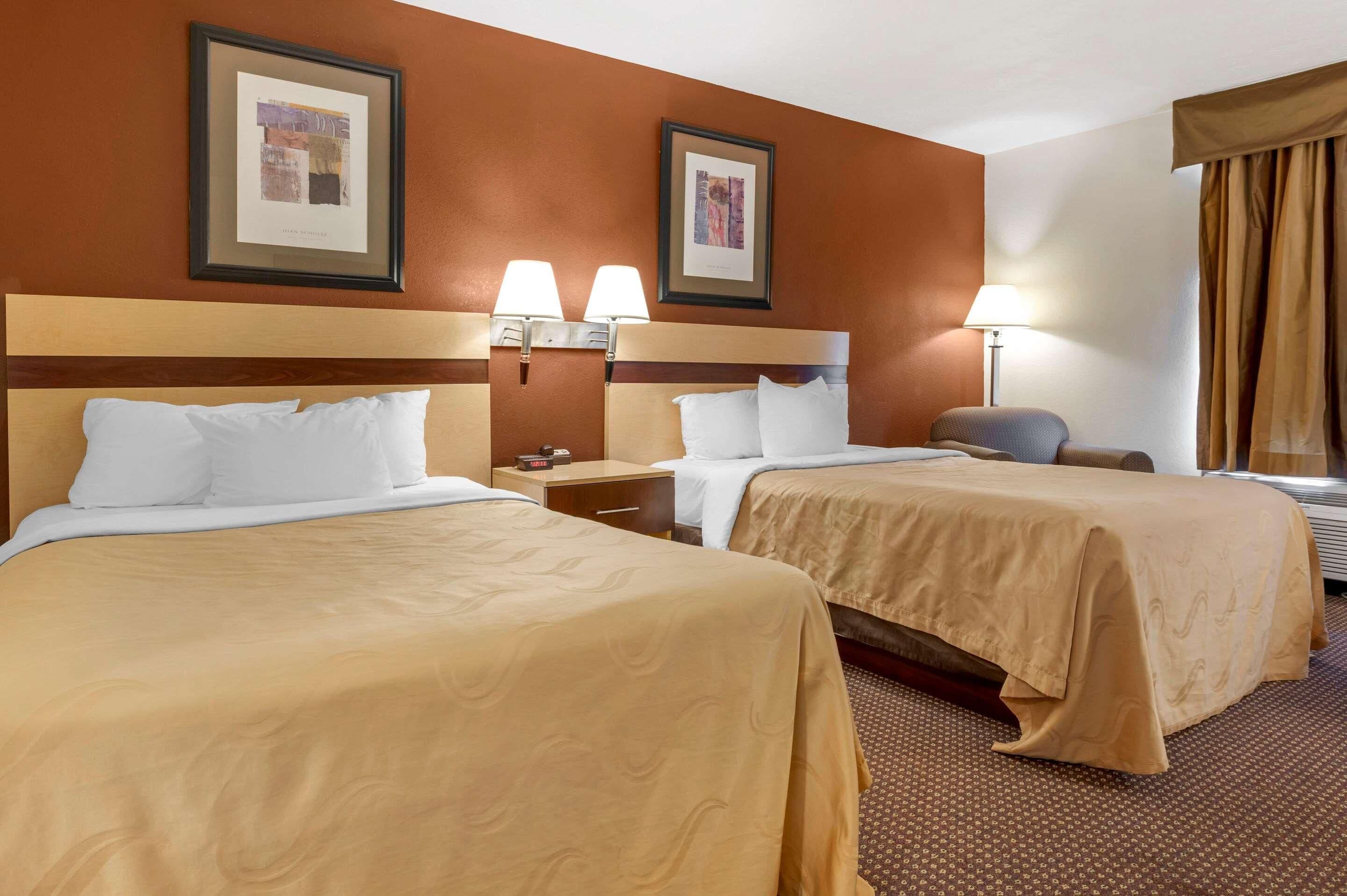 A hotel room with two beds, a nightstand, a lamp, and framed pictures on the wall.