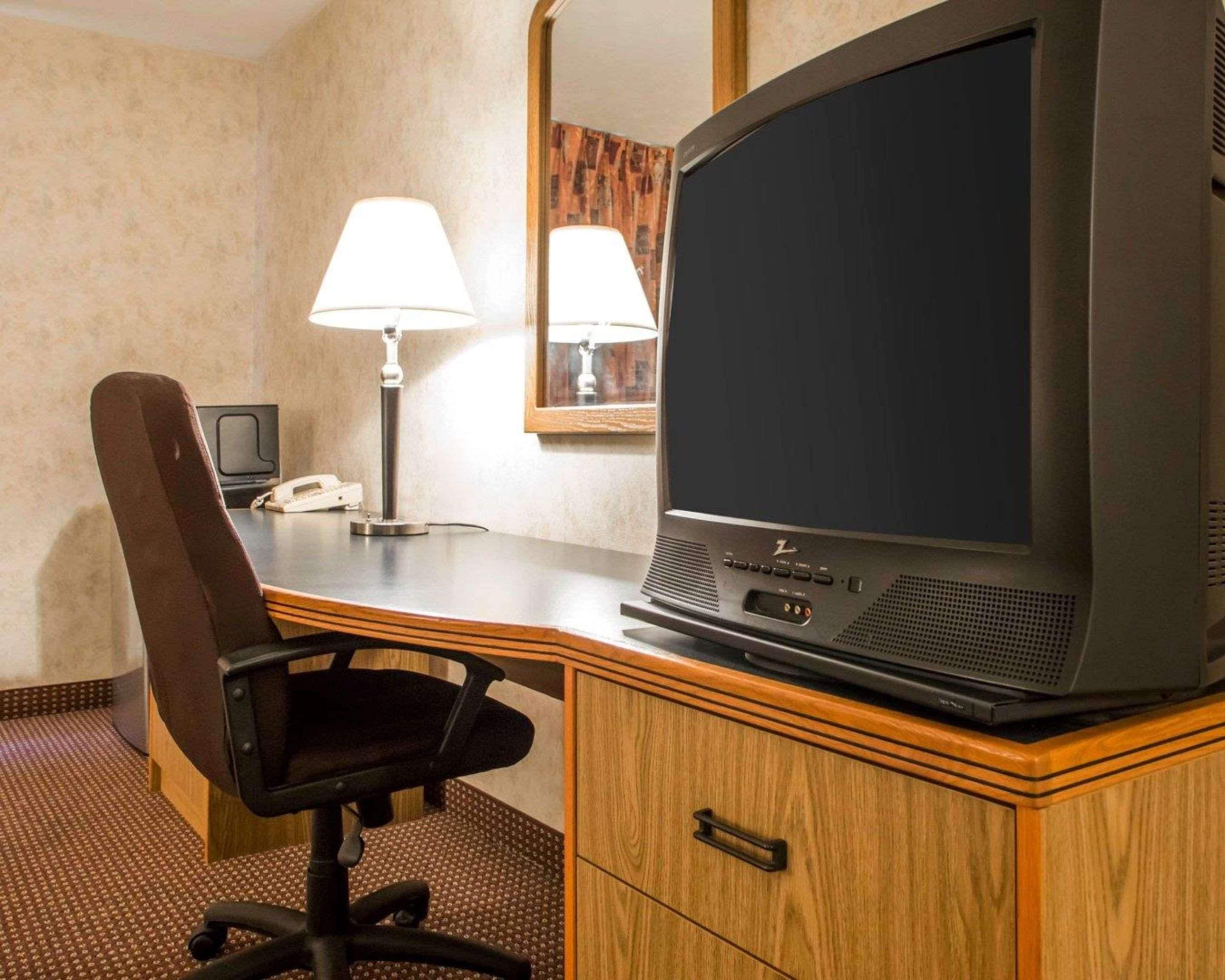 A hotel room with a wooden desk, a black office chair, a television, a telephone, and a lamp.