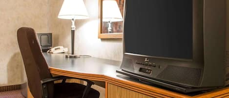 A hotel room with a wooden desk, a black office chair, a television, a telephone, and a lamp.