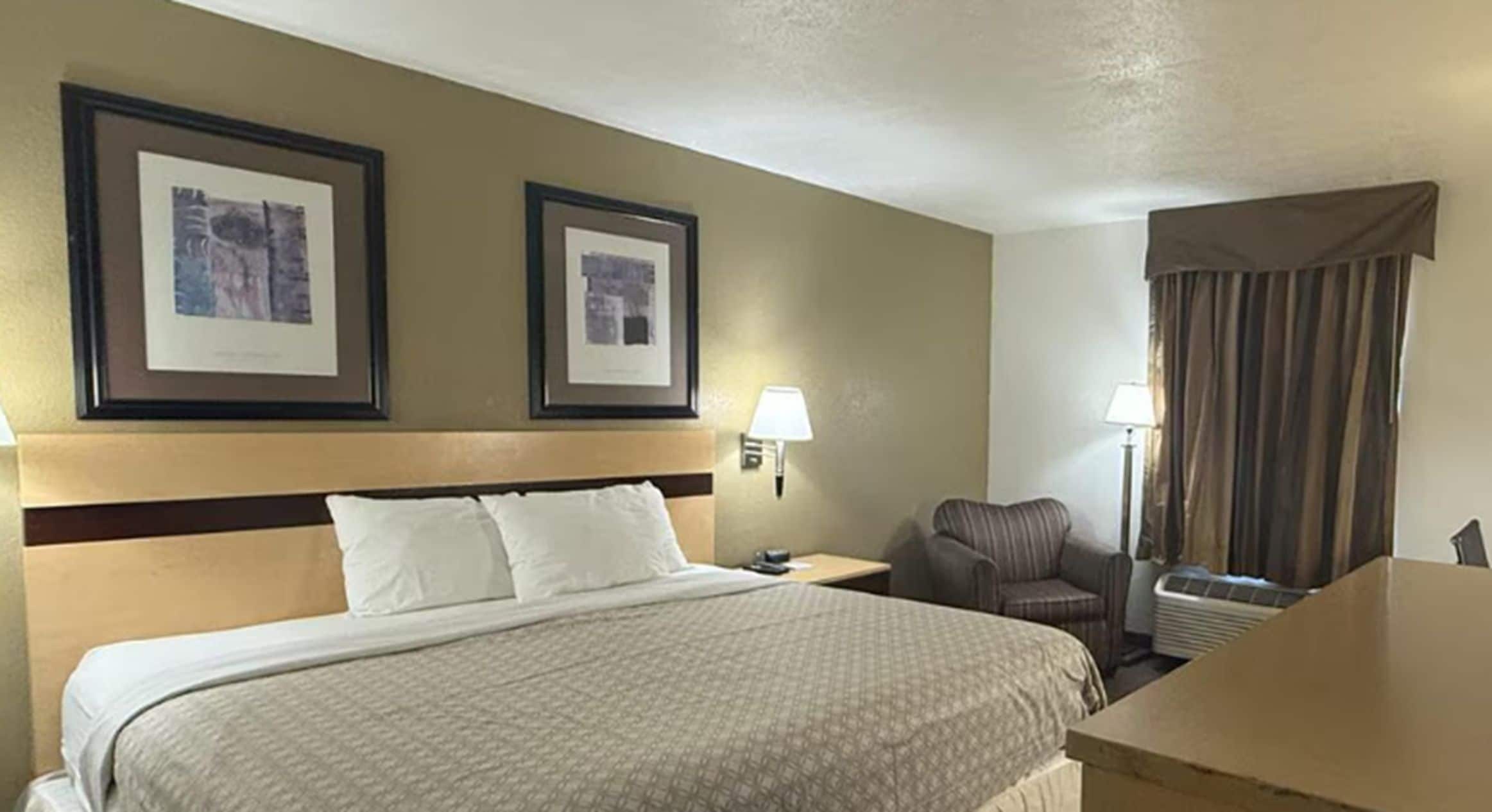 A hotel room with a bed, two framed pictures on the wall, a chair, a desk, and a window with curtains.