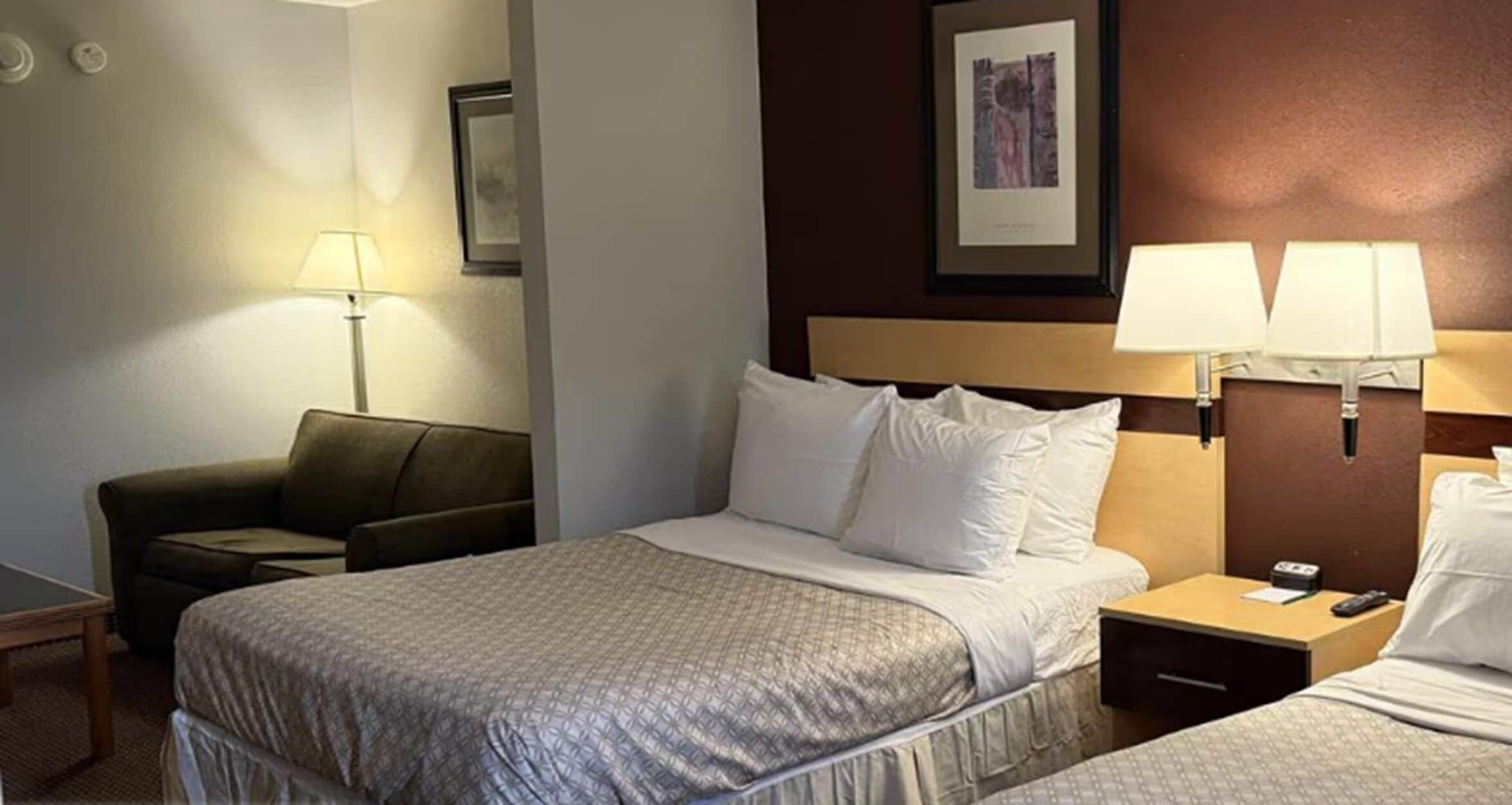 Hotellihuone, jossa on sänky, kaksi yöpöytälamppua, yöpöytä, tuoli ja seinällä oleva kehystetty kuva.