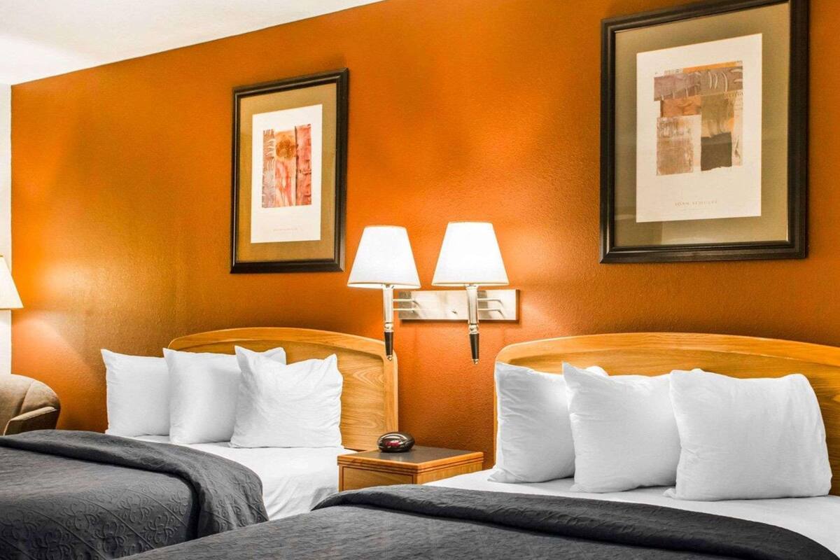 A hotel room with two beds, each with white linens and a dark gray bedspread. There are two wall-mounted lamps on either side of the beds, and two framed artworks above the beds.