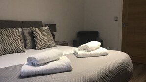 Een opgemaakt bed met twee opgerolde handdoeken, een hoofdbord met gestreepte kussens, een nachtkastje en een stoel.