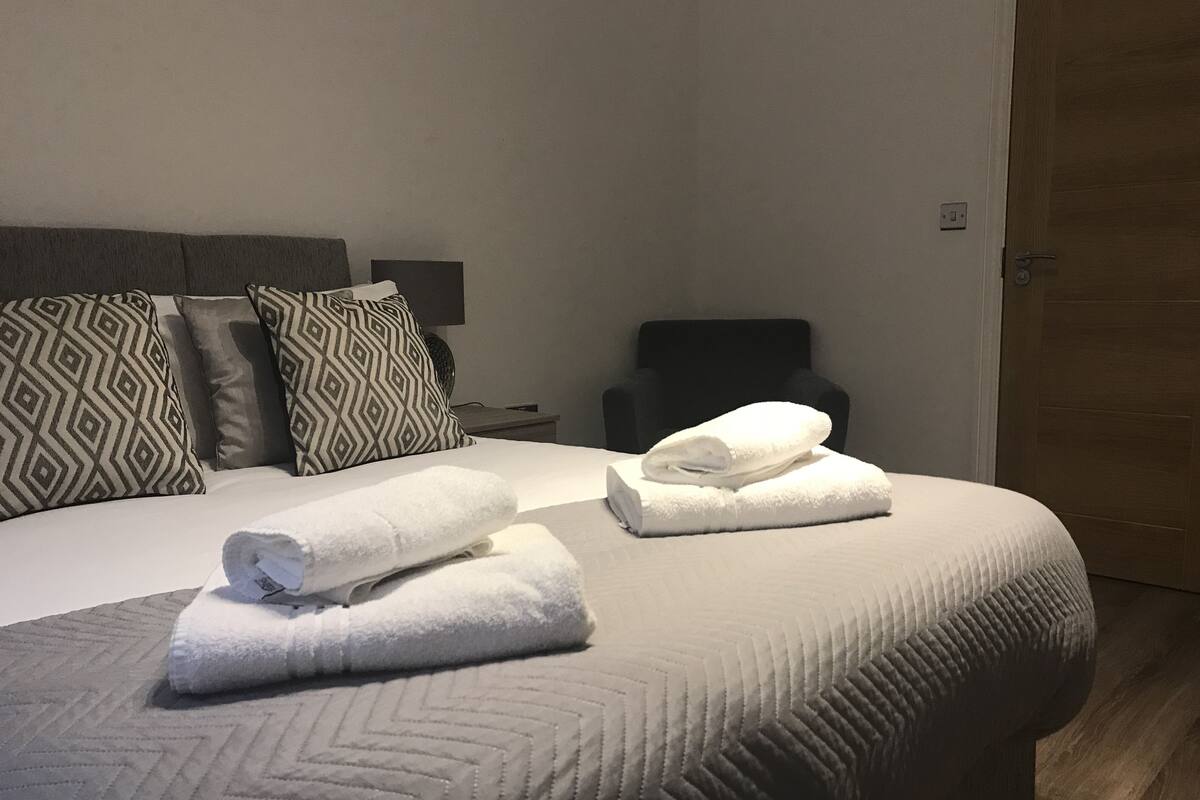 Una cama bien tendida con dos toallas enrolladas, un cabecero con almohadones estampados, una mesita de noche y una silla.