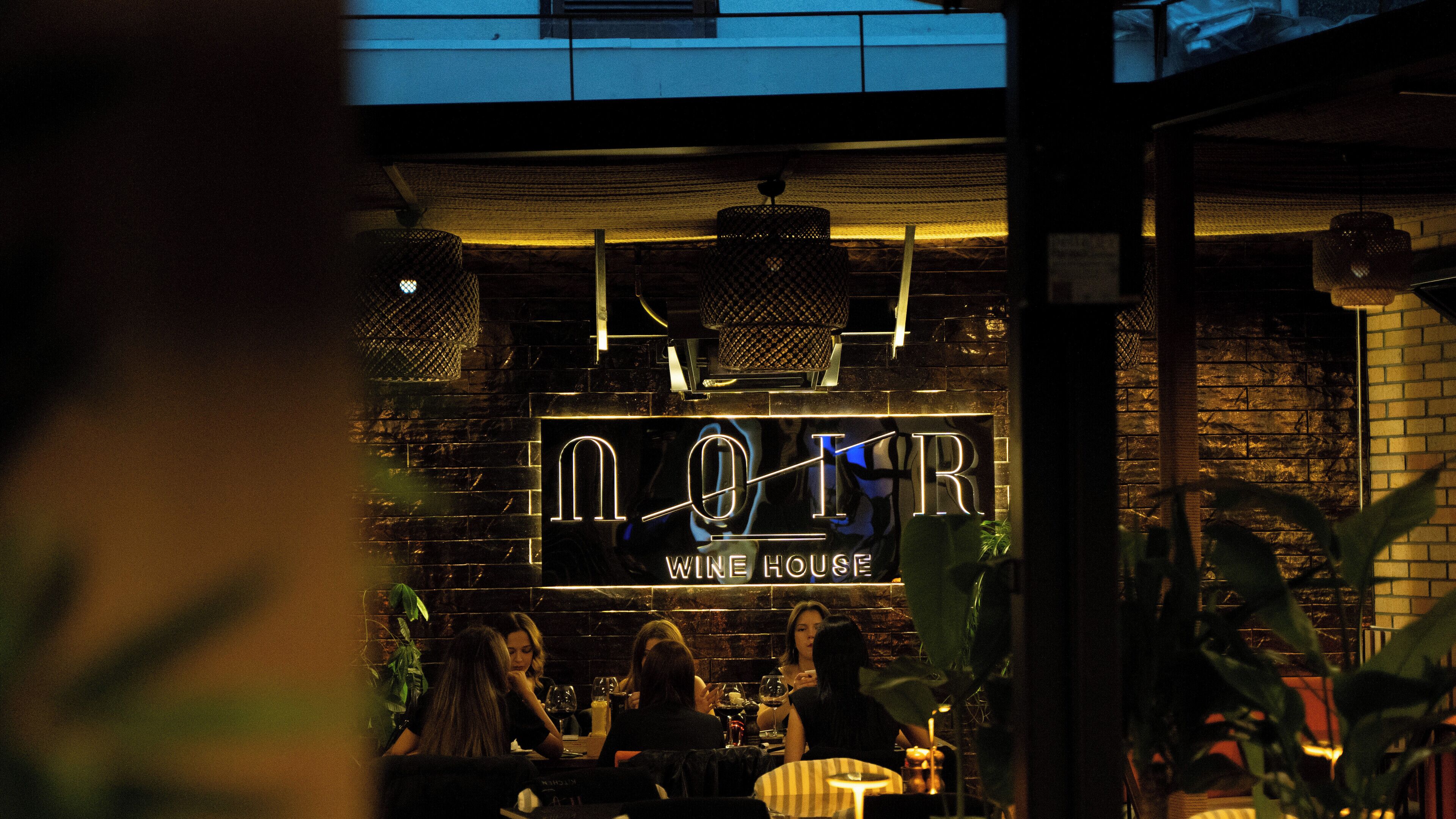A wine house interior with a brick wall, hanging lights, and a group of people seated at tables.
