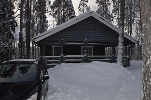 A log cabin with a snow-covered roof and a wooden deck, surrounded by tall trees. - Cottage H62 Ruokolahti (Ruokolahti)