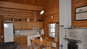 A log cabin interior with a dining table, chairs, wooden cabinets, a kitchen area with appliances, and a fireplace. - Cottage H62 Ruokolahti (Ruokolahti)